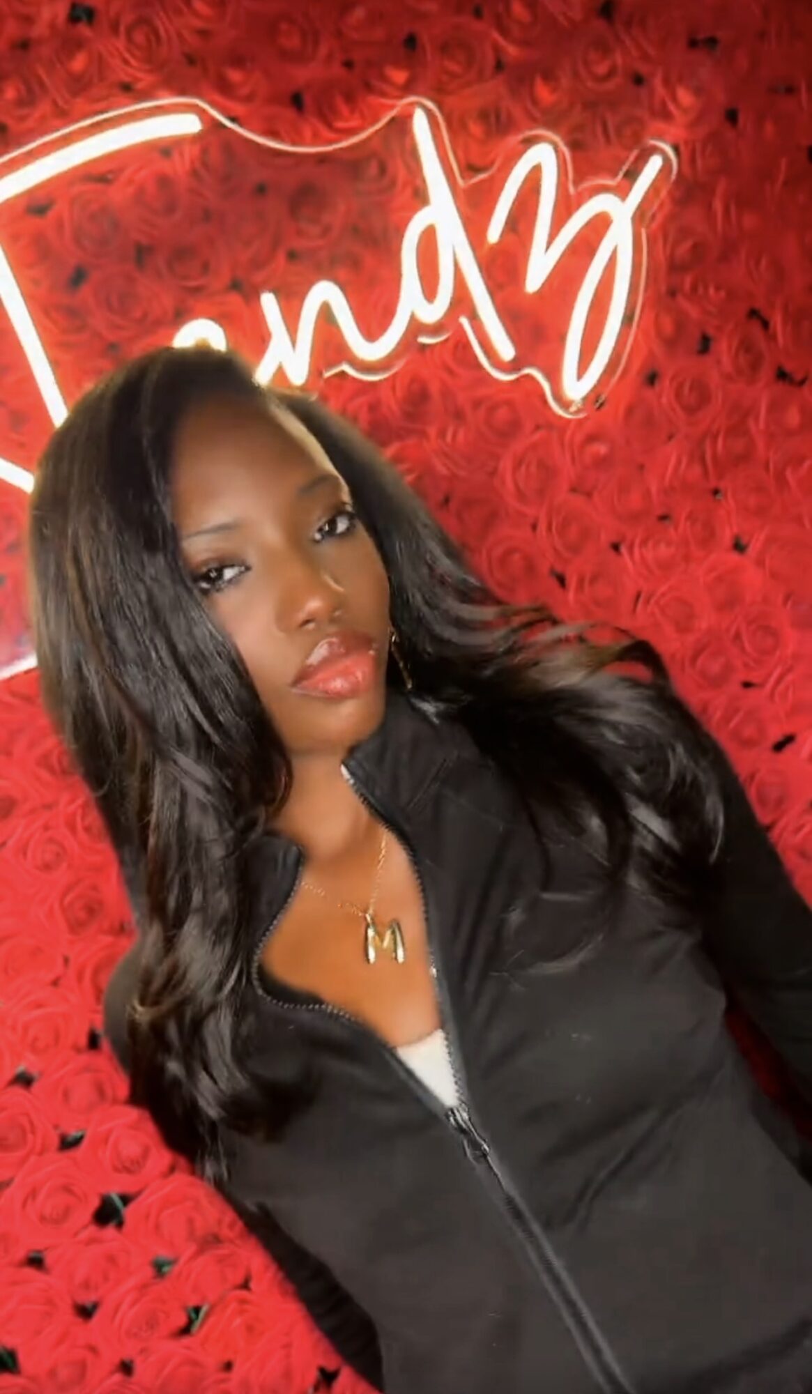 Young woman with long wavy hair, wearing a black jacket, standing in front of a red background with neon sign.