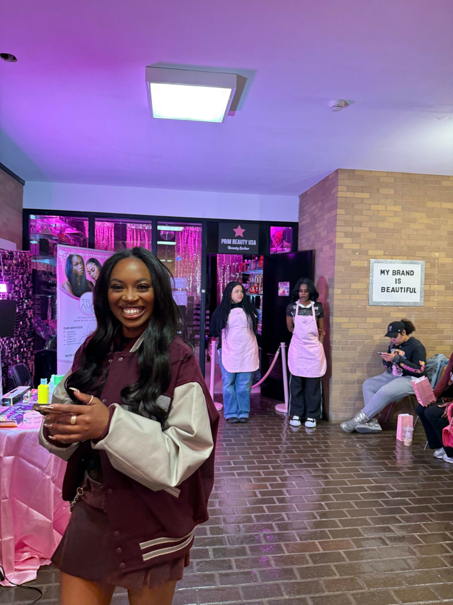 Woman smiling and holding a phone, standing in front of a pink-lit display with people sitting nearby.