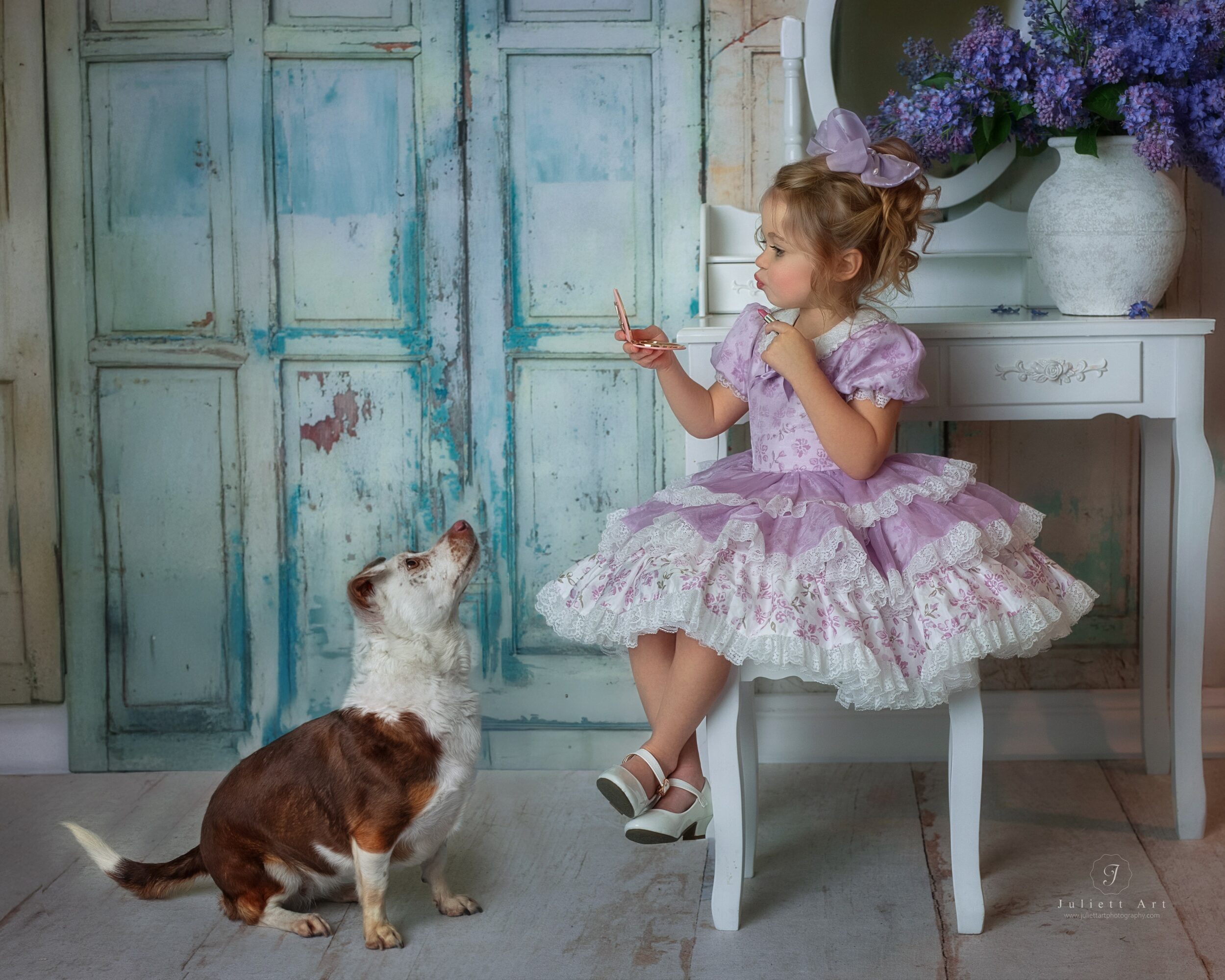 Young girl in a purple dress sitting on a white chair, holding a flower, with a dog looking up at her, in a room with a blue door and purple flowers. Word count: 27