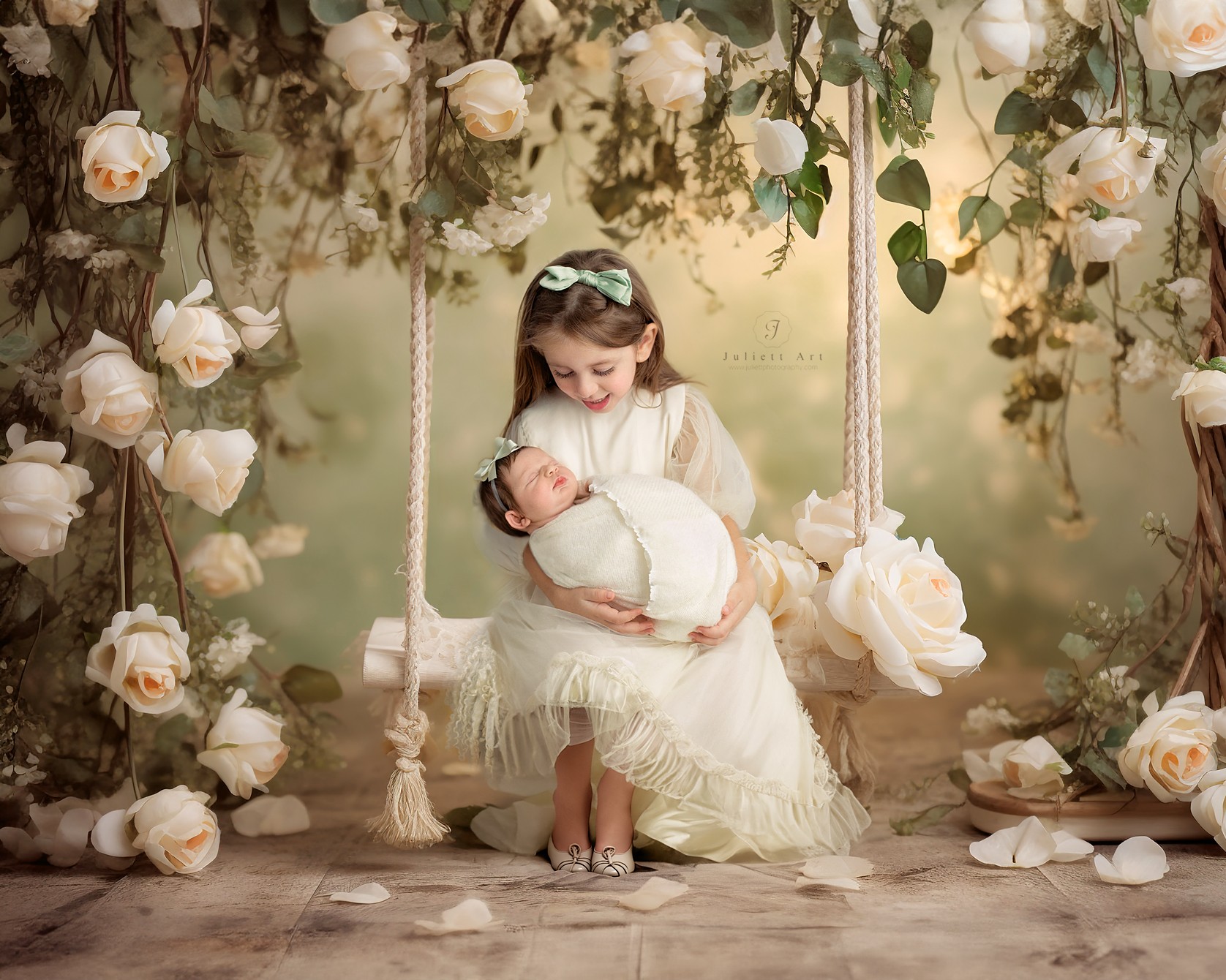 A young girl in a white dress holding a baby on a swing surrounded by white roses and greenery.