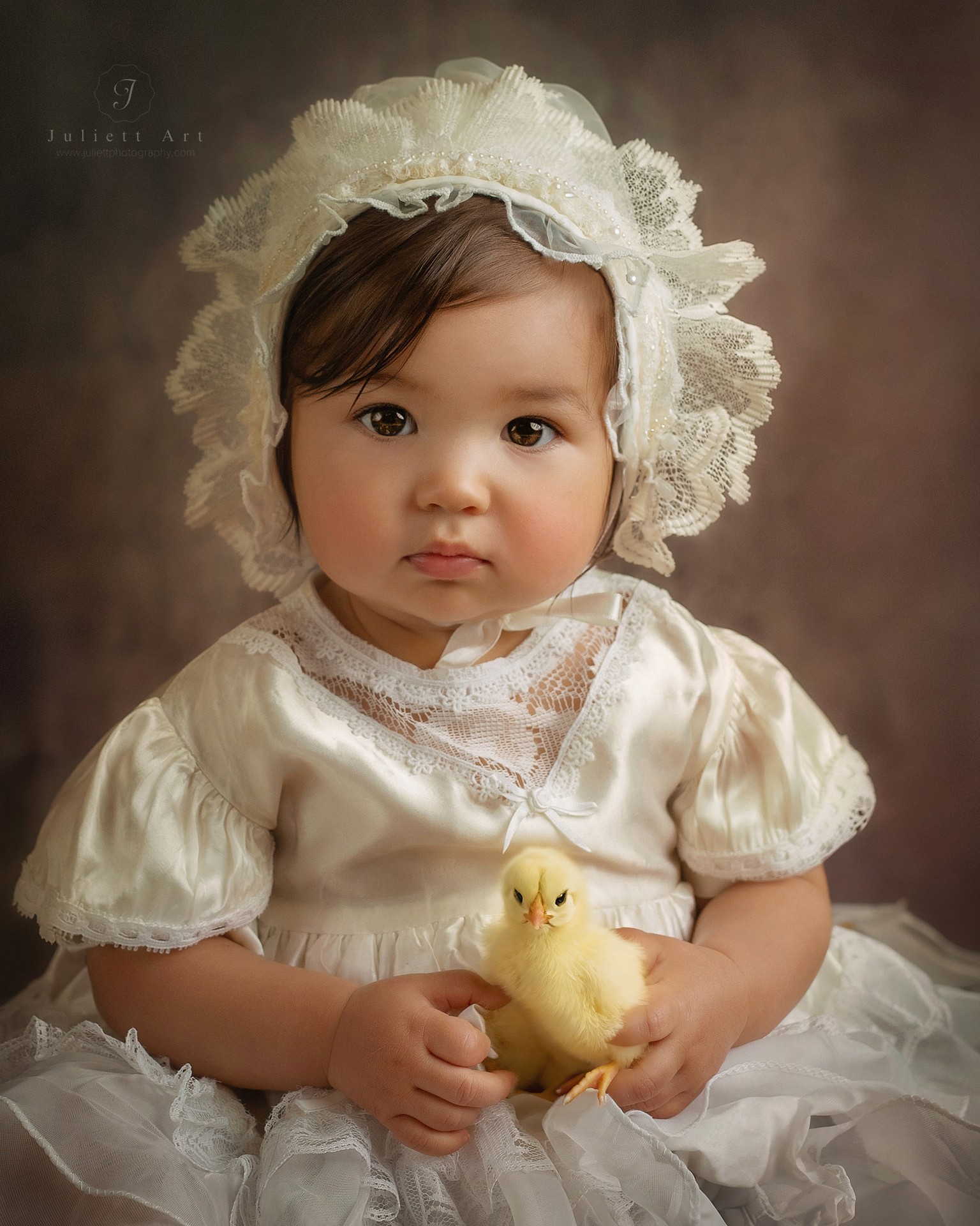 Child in vintage dress holding a yellow chick, wearing a lace bonnet, against a brown background.