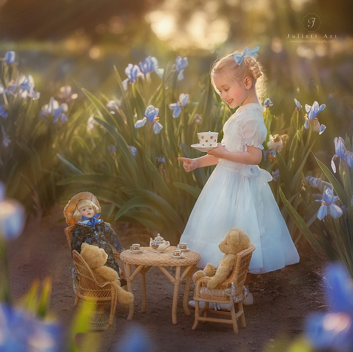 Young girl in a white dress holding a teacup among blue flowers, with a doll and miniature furniture nearby.