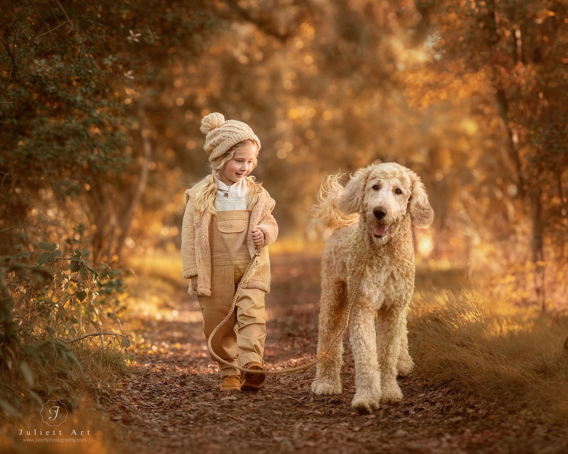 A young girl in warm clothing walks a dog on a leash through an autumn forest path.