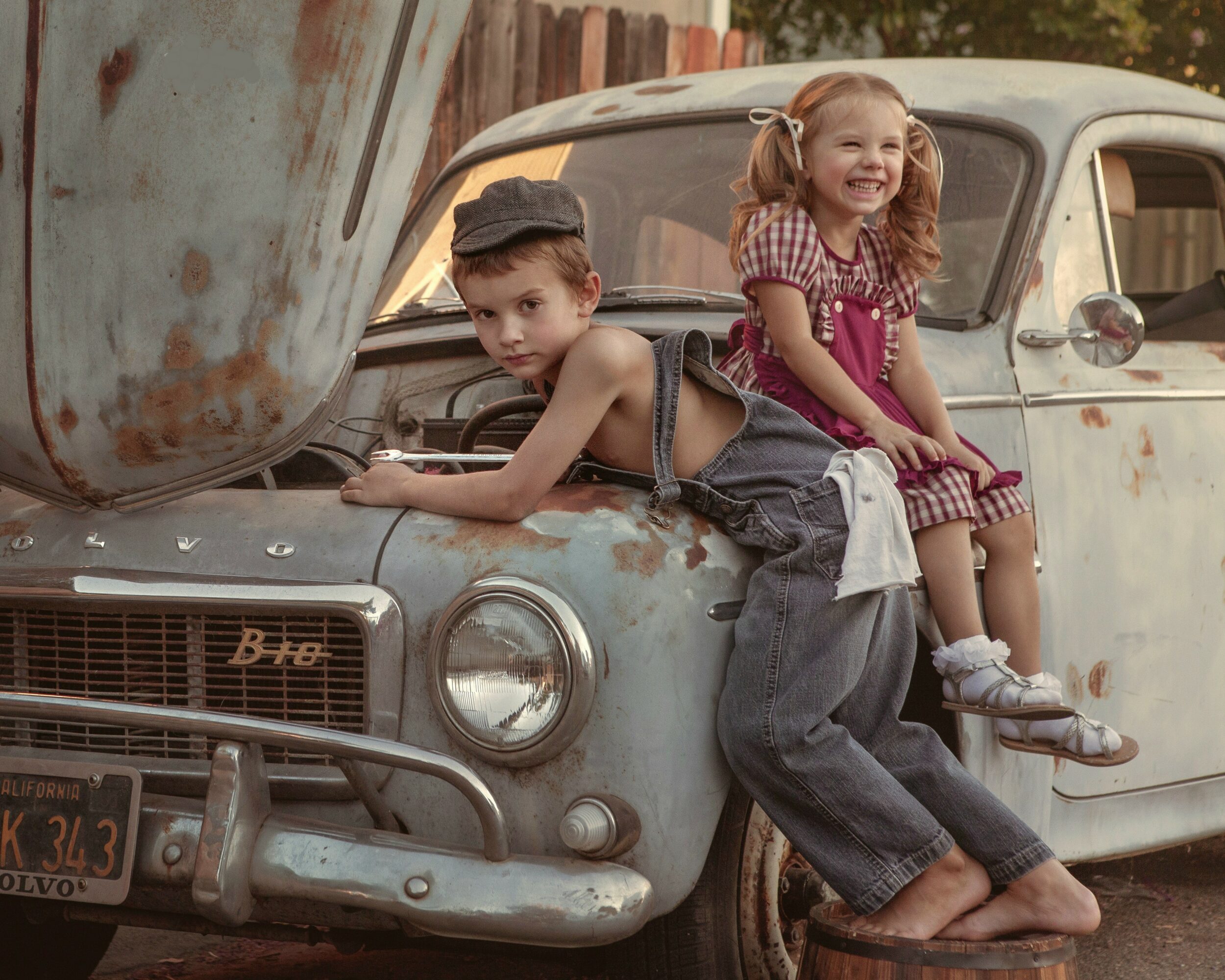 Two children sit on the hood of an old, rusty car, smiling and playing outdoors.