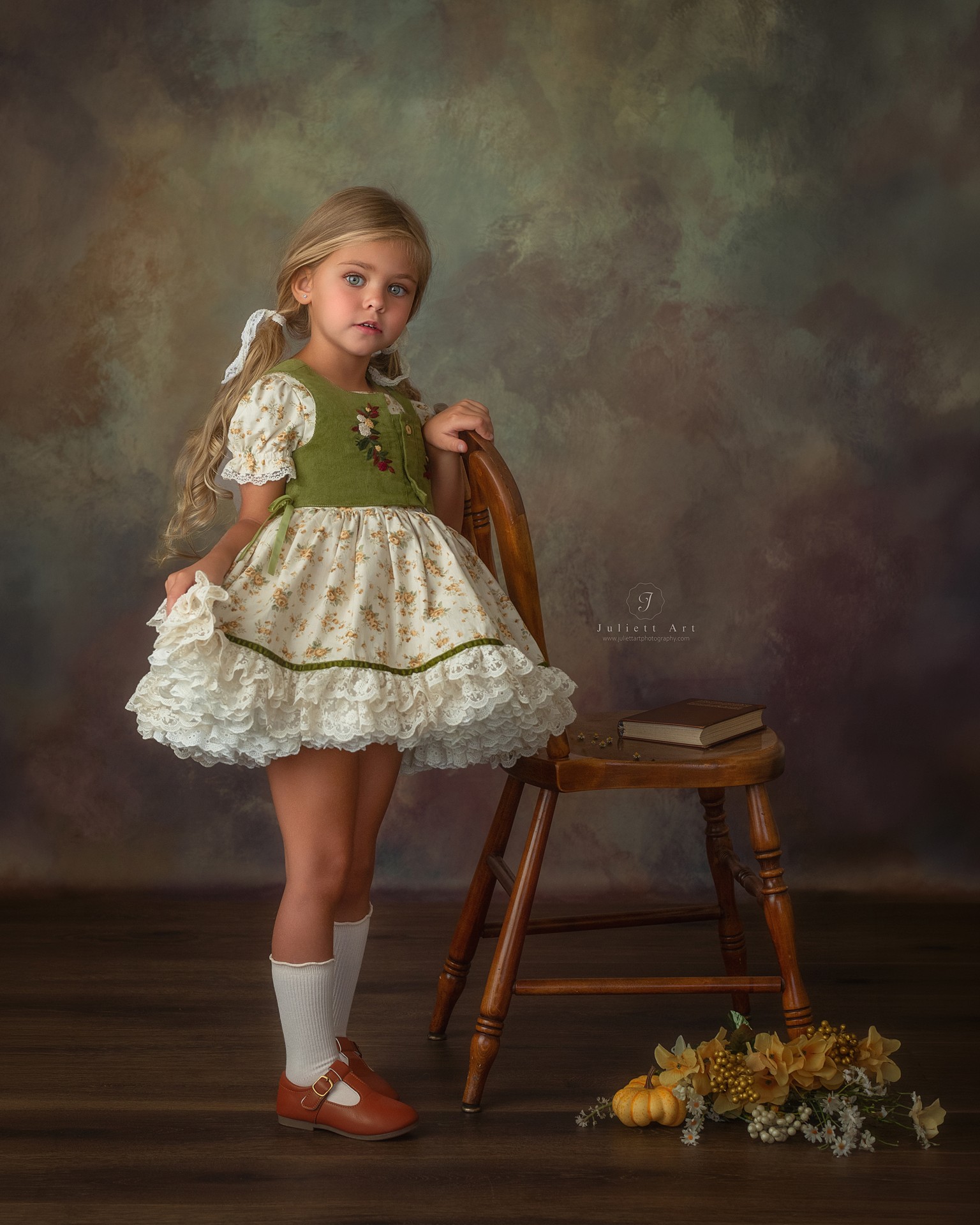 Young girl in a vintage dress standing next to a wooden chair with books and flowers on the floor.