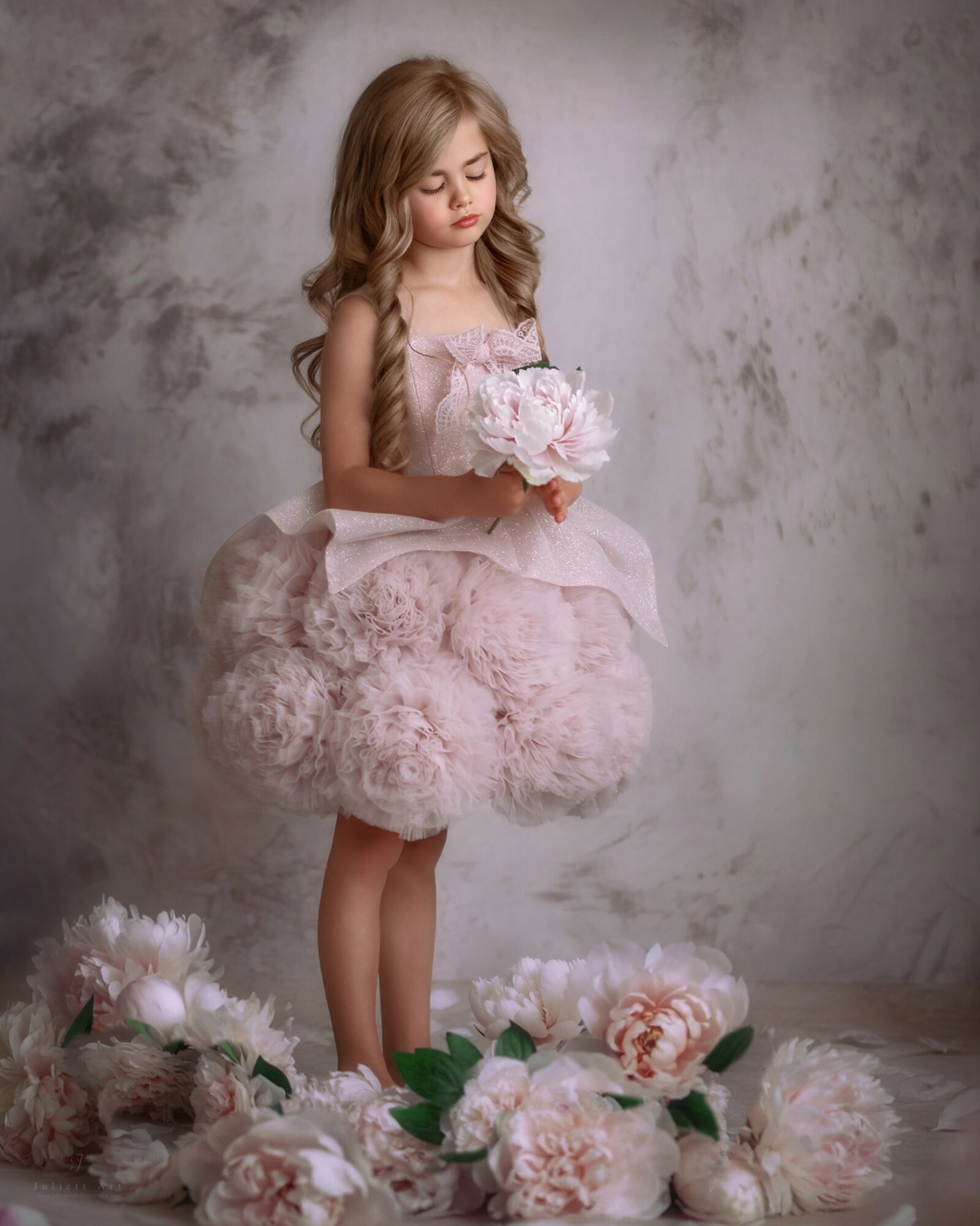 Young girl in a pink dress holding a bouquet of flowers, standing among large flowers on the floor, neutral background.