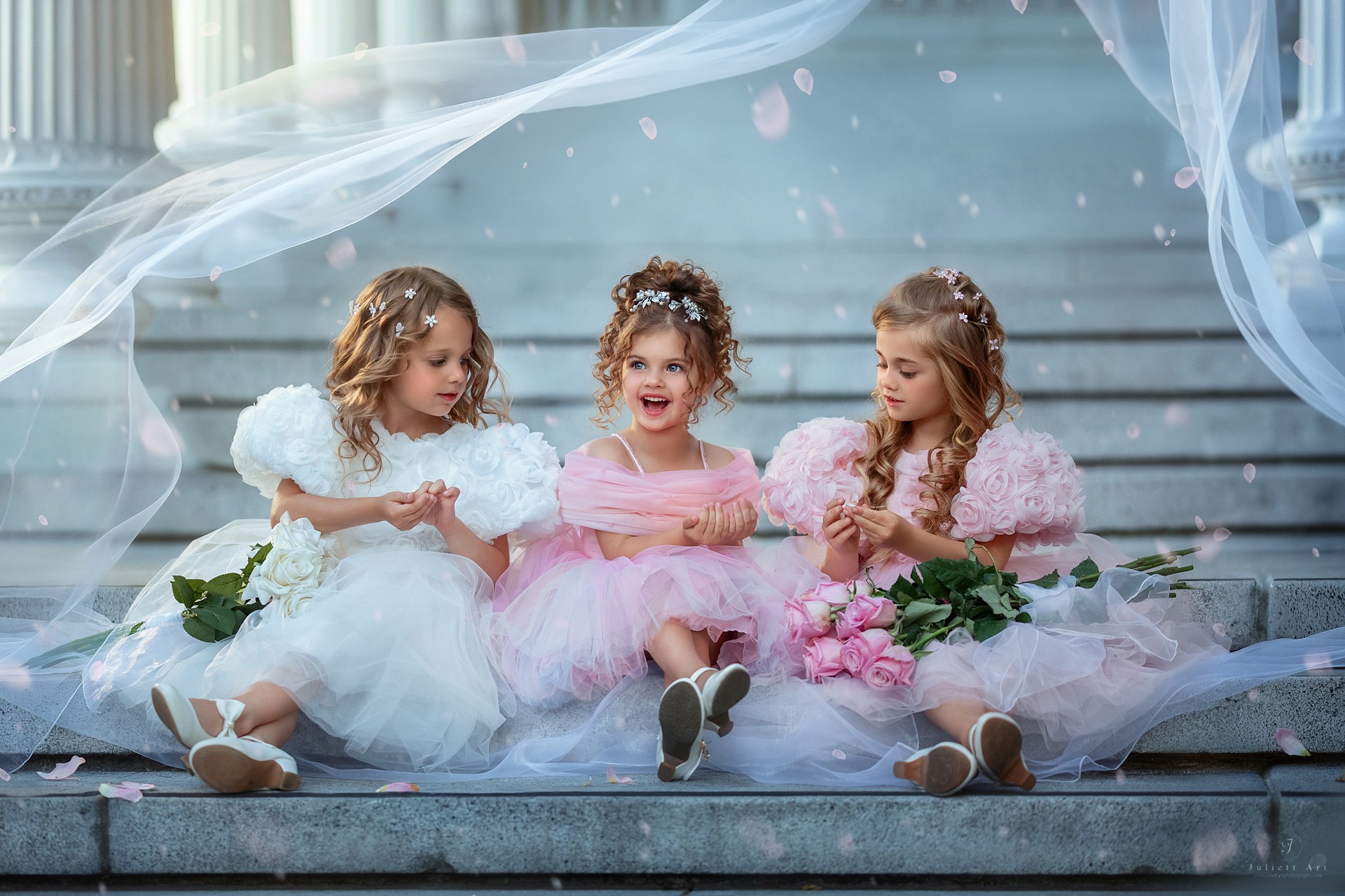 Three young girls in fancy dresses sitting on steps, holding flowers, with decorative fabric and flowers around them.
