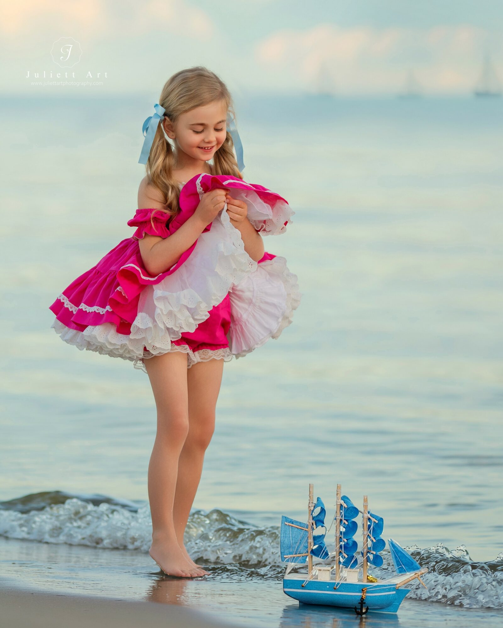 Young girl in pink dress playing with a toy sailboat on the beach, smiling, ocean in background.