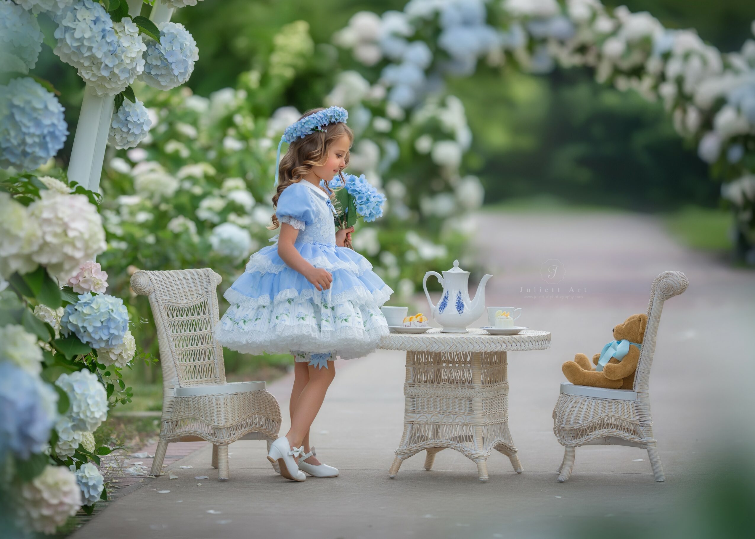 A young girl in a blue dress and headband stands at a small table with teapots, surrounded by white flowers and garden chairs.