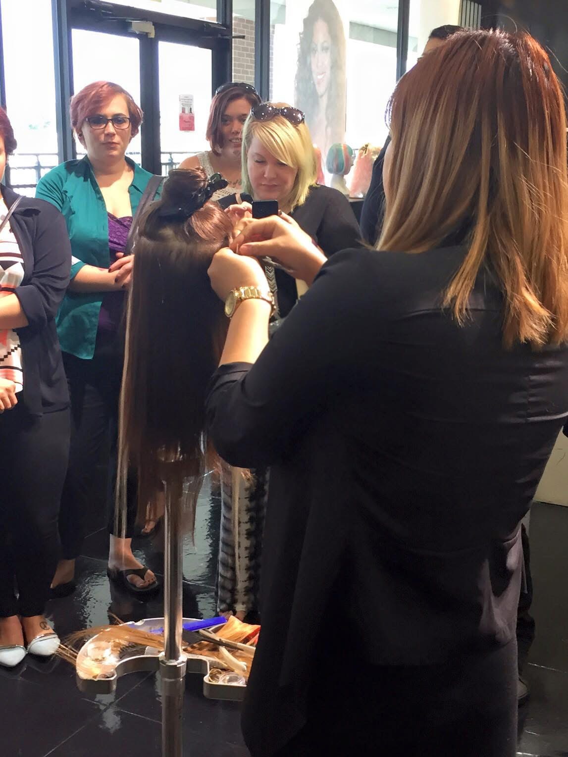 Hairdresser demonstrating hair styling to a group of women in a salon, with hair tools and a mannequin head present.