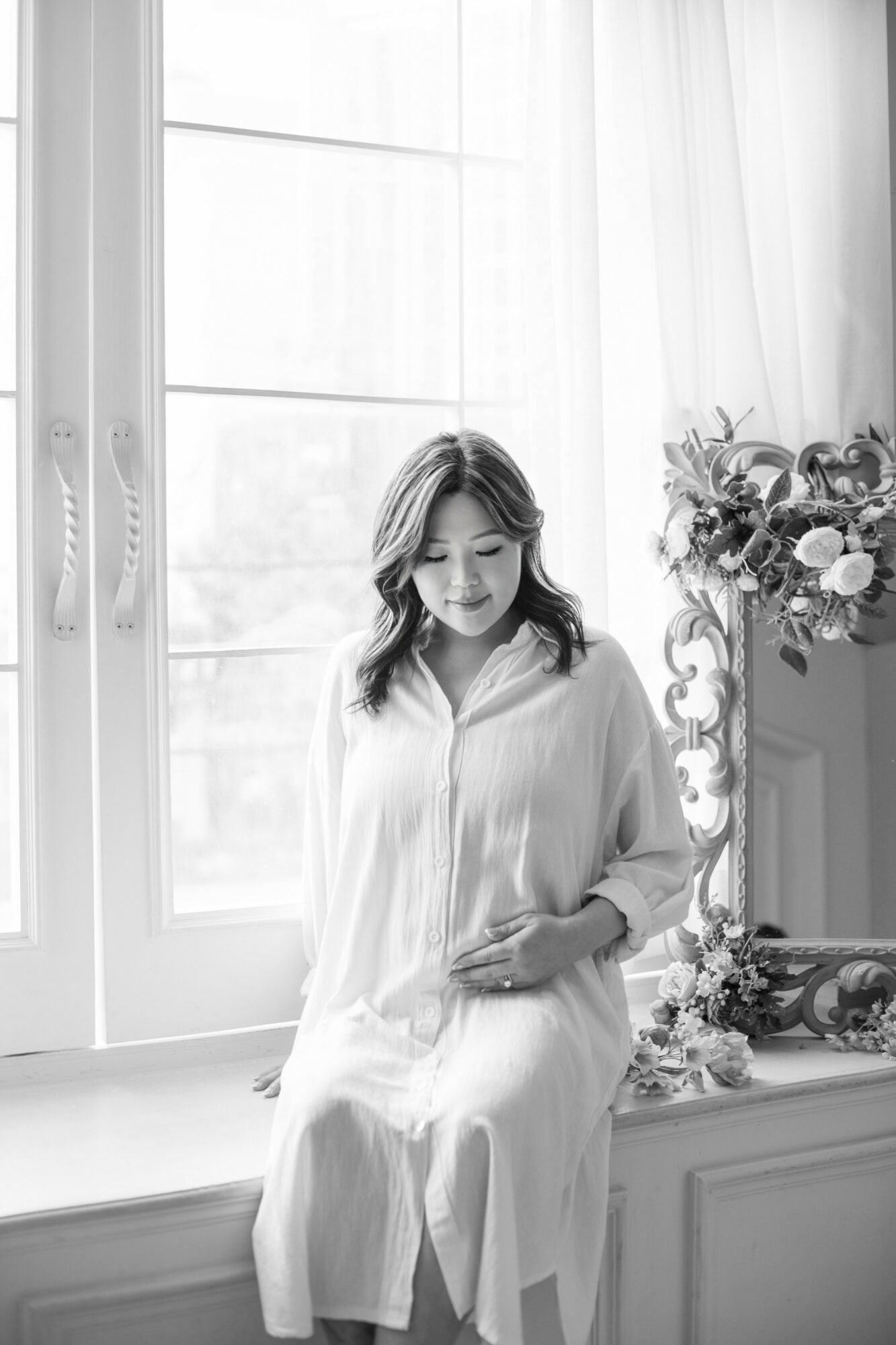 Woman sitting on windowsill, smiling, with a flower arrangement nearby, in a bright room with large windows.