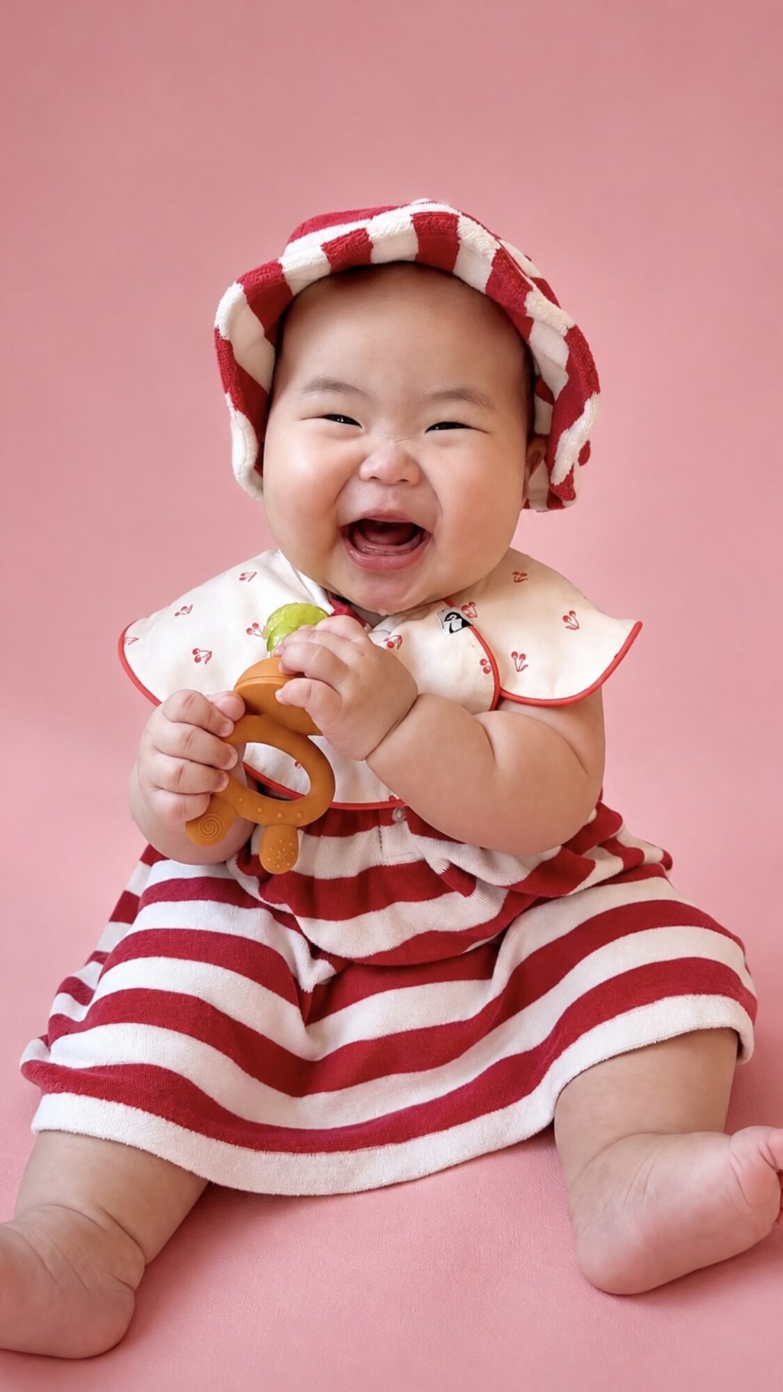 Smiling baby wearing a striped dress and hat, holding a toy, sitting against a pink background.