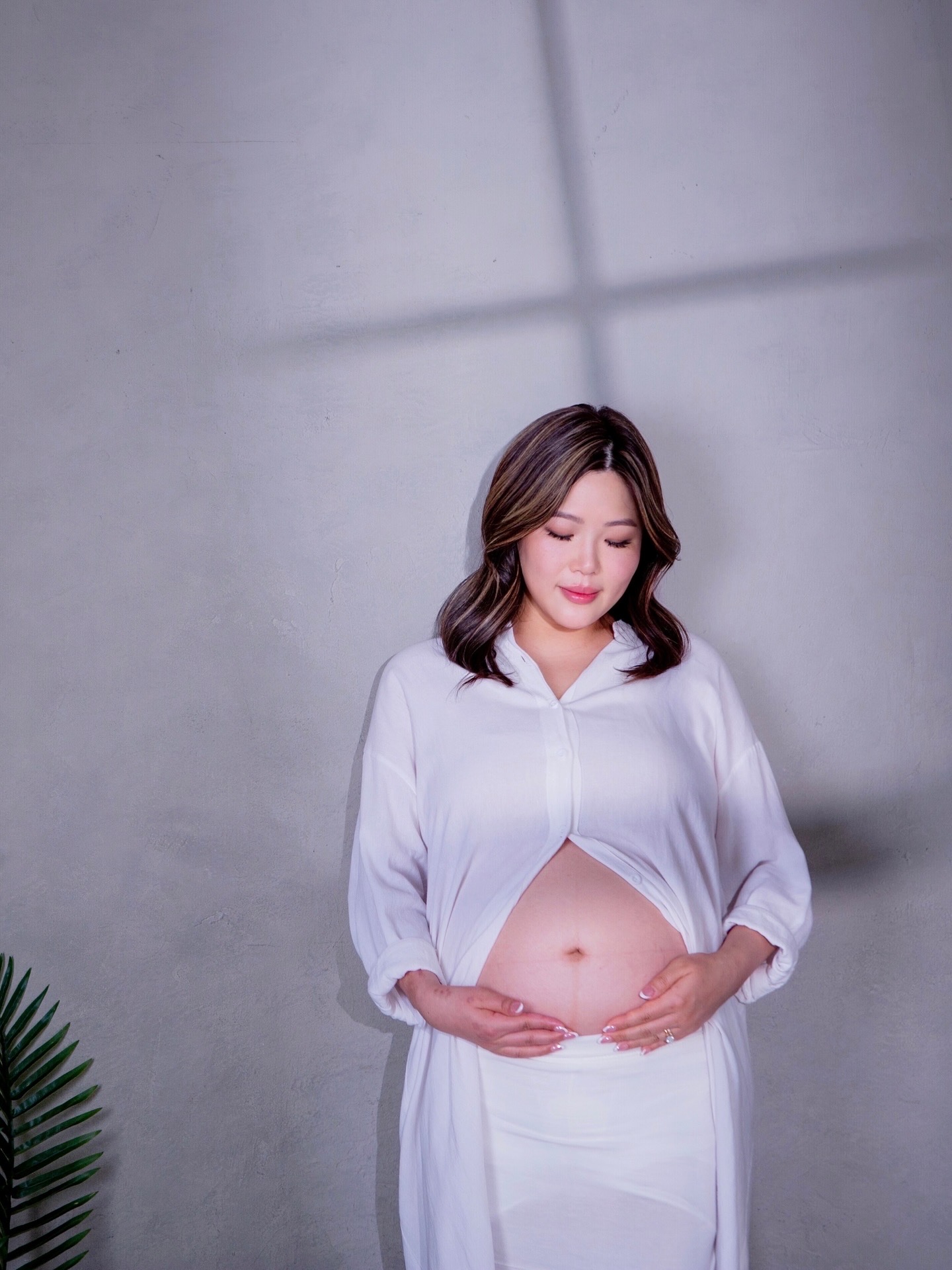 Pregnant woman with hands on belly, standing against a plain wall, looking down, wearing a white outfit.