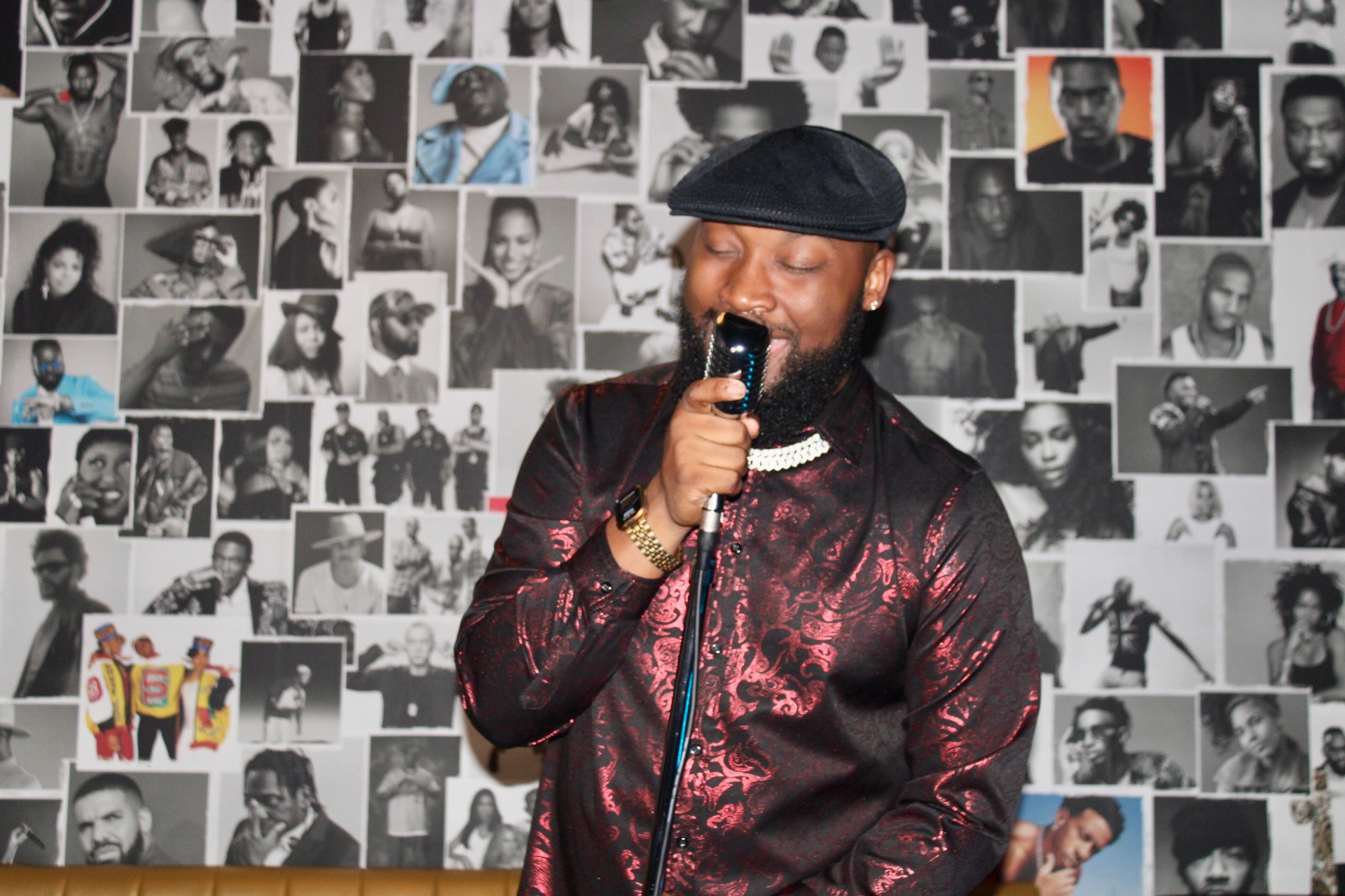 Man with a beard wearing a black beret and patterned jacket, holding a microphone, standing in front of a collage of black-and-white photos.