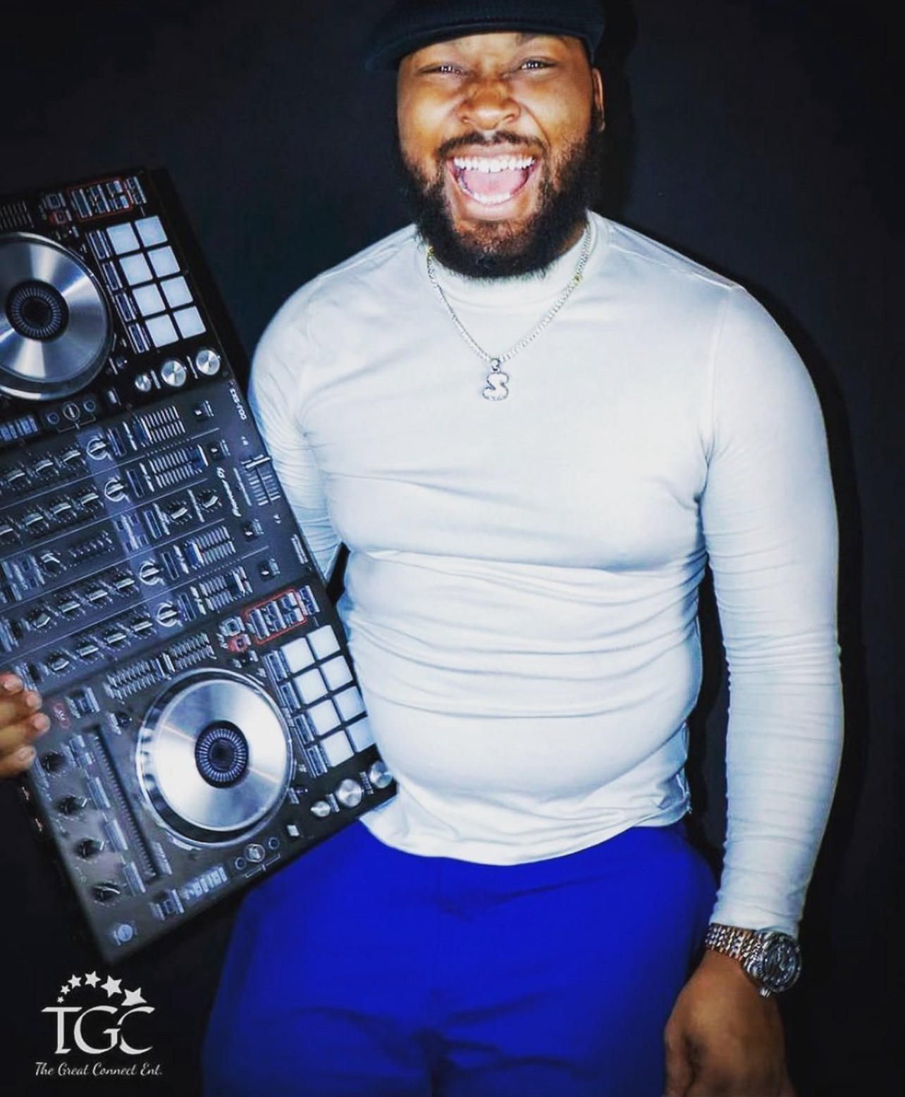 Person smiling, holding DJ equipment, wearing a white long-sleeve shirt, blue pants, and a black hat.