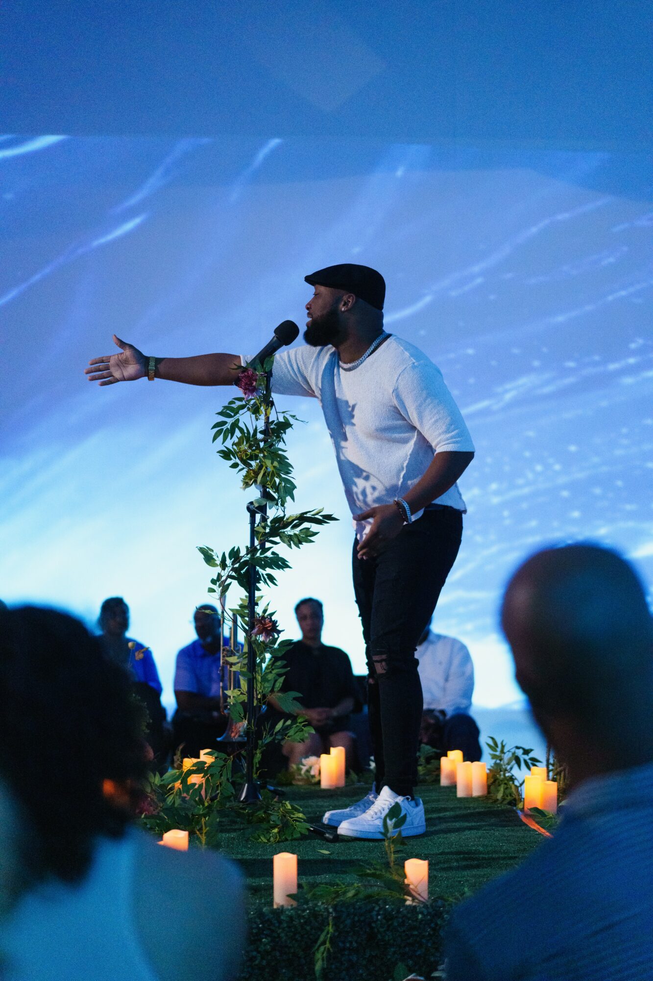 Performer on stage with a microphone, gesturing with one arm, surrounded by candles and audience, blue background.
