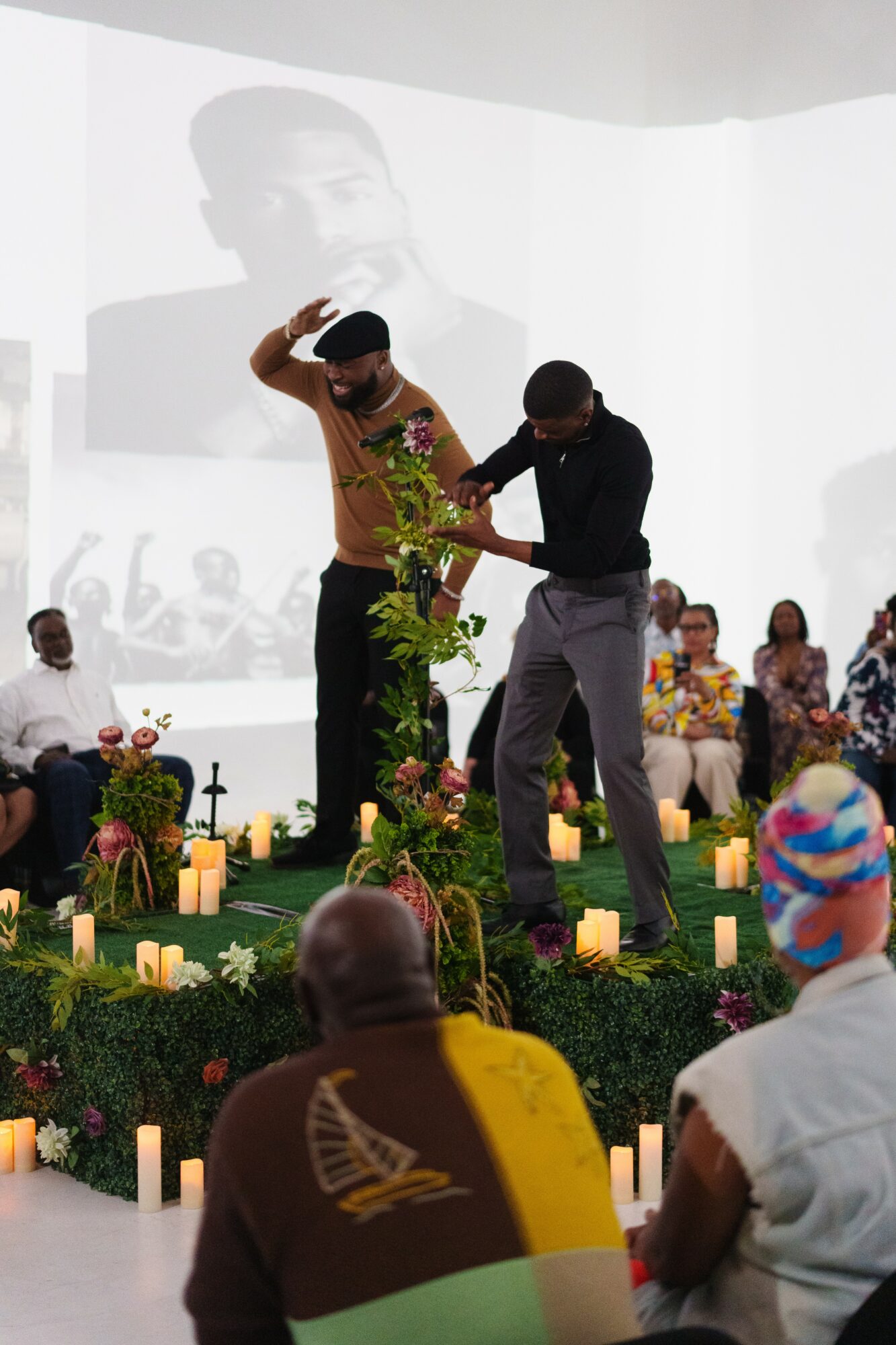 Two men on stage, one with a hat, exchanging a plant, with seated audience and candles in the background.