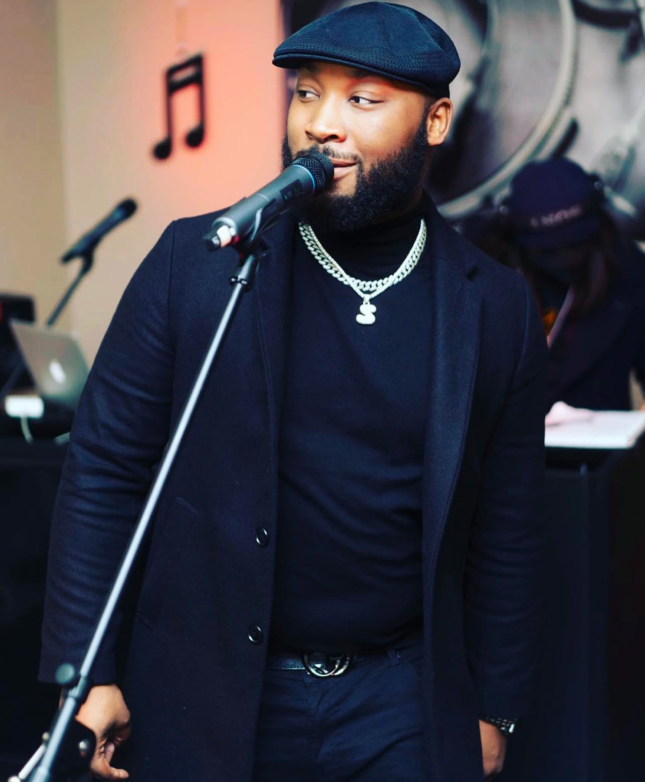 Man with beard and cap singing into microphone, wearing a chain necklace, in a music studio or performance space.