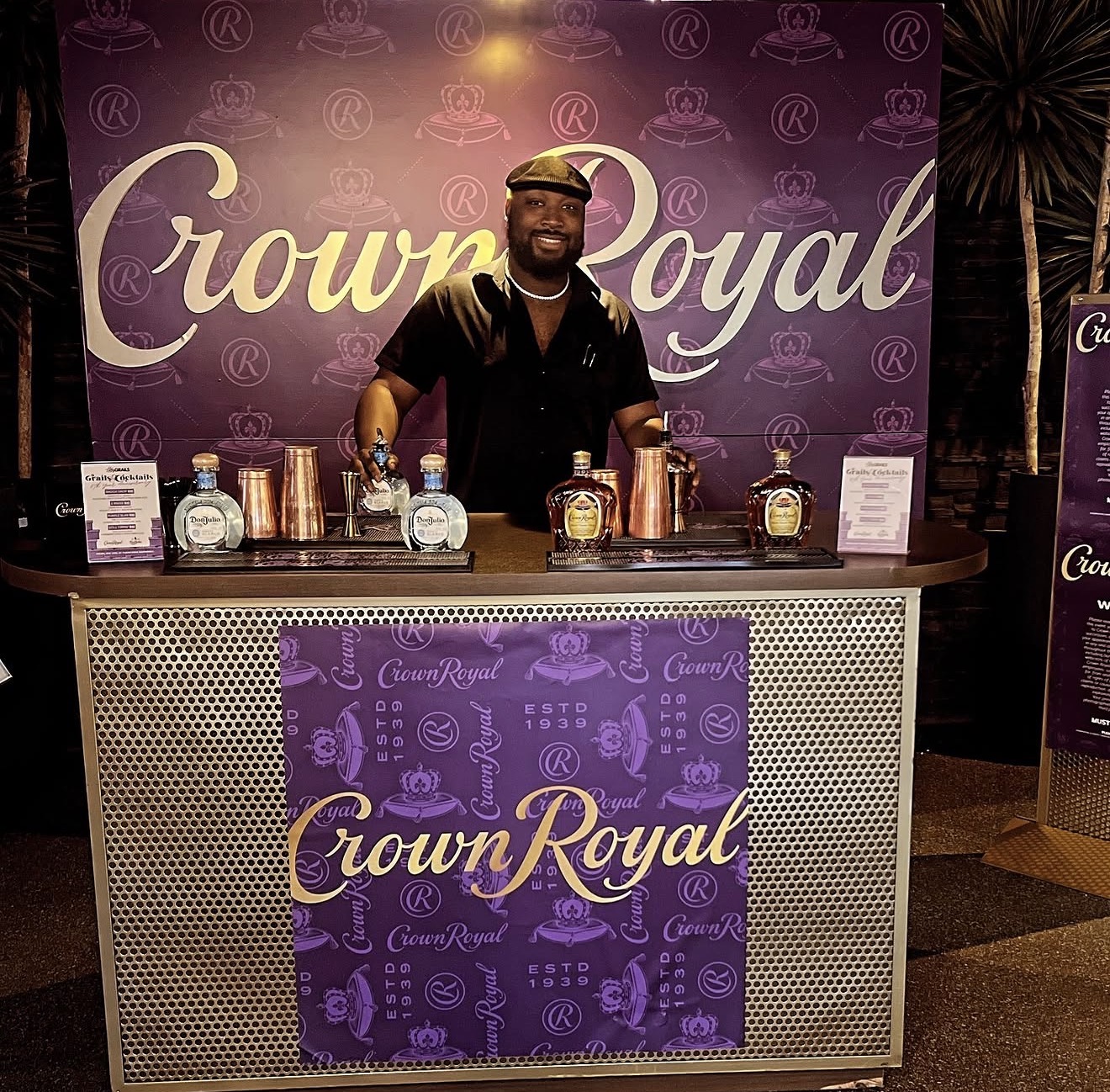 Person standing behind a Crown Royal branded bar setup with bottles and cups, purple background, nighttime setting.