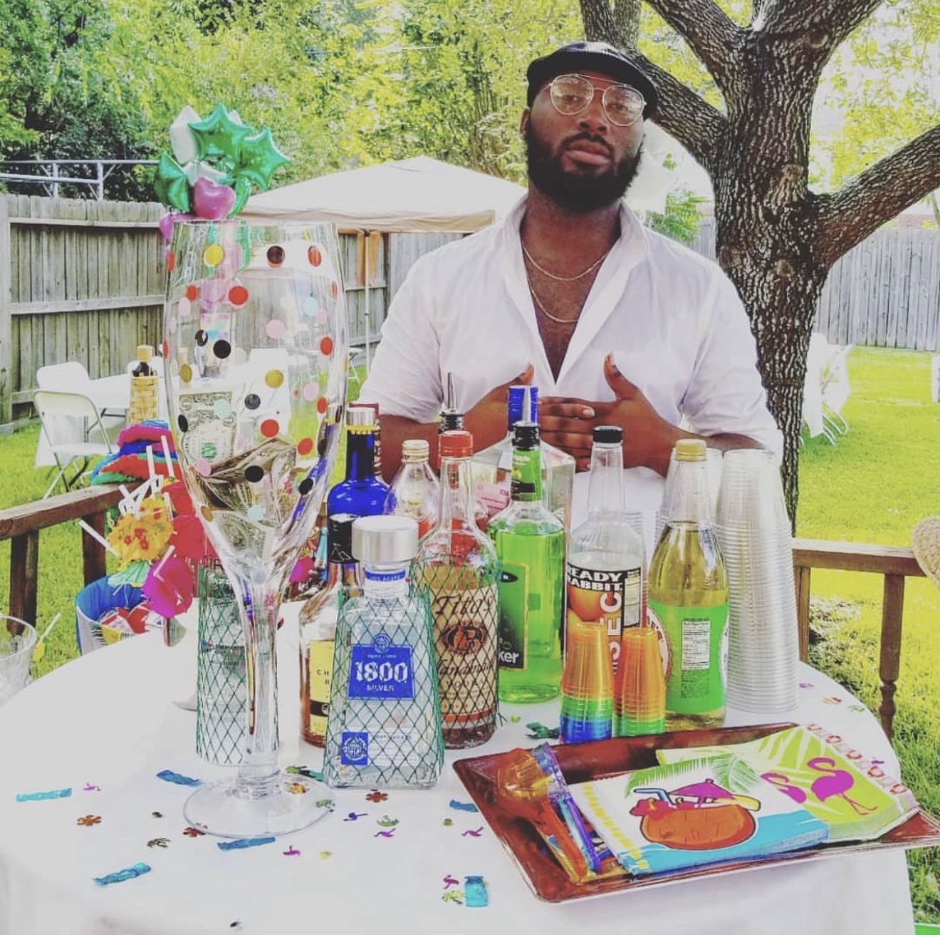 Man standing behind a table with drinks and party decorations outdoors, trees and a fence in the background.