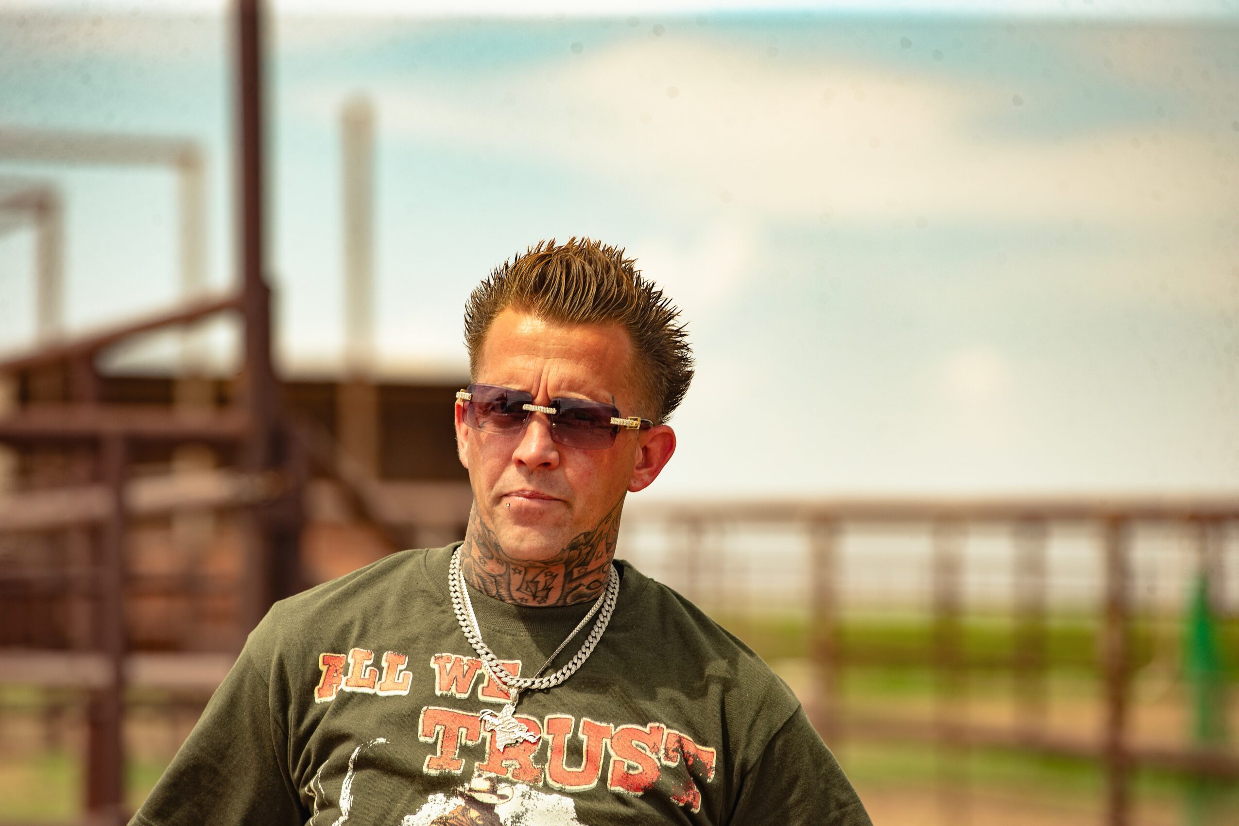Man with sunglasses, short hair, and tattoos wearing a T-shirt and chain necklace outdoors.