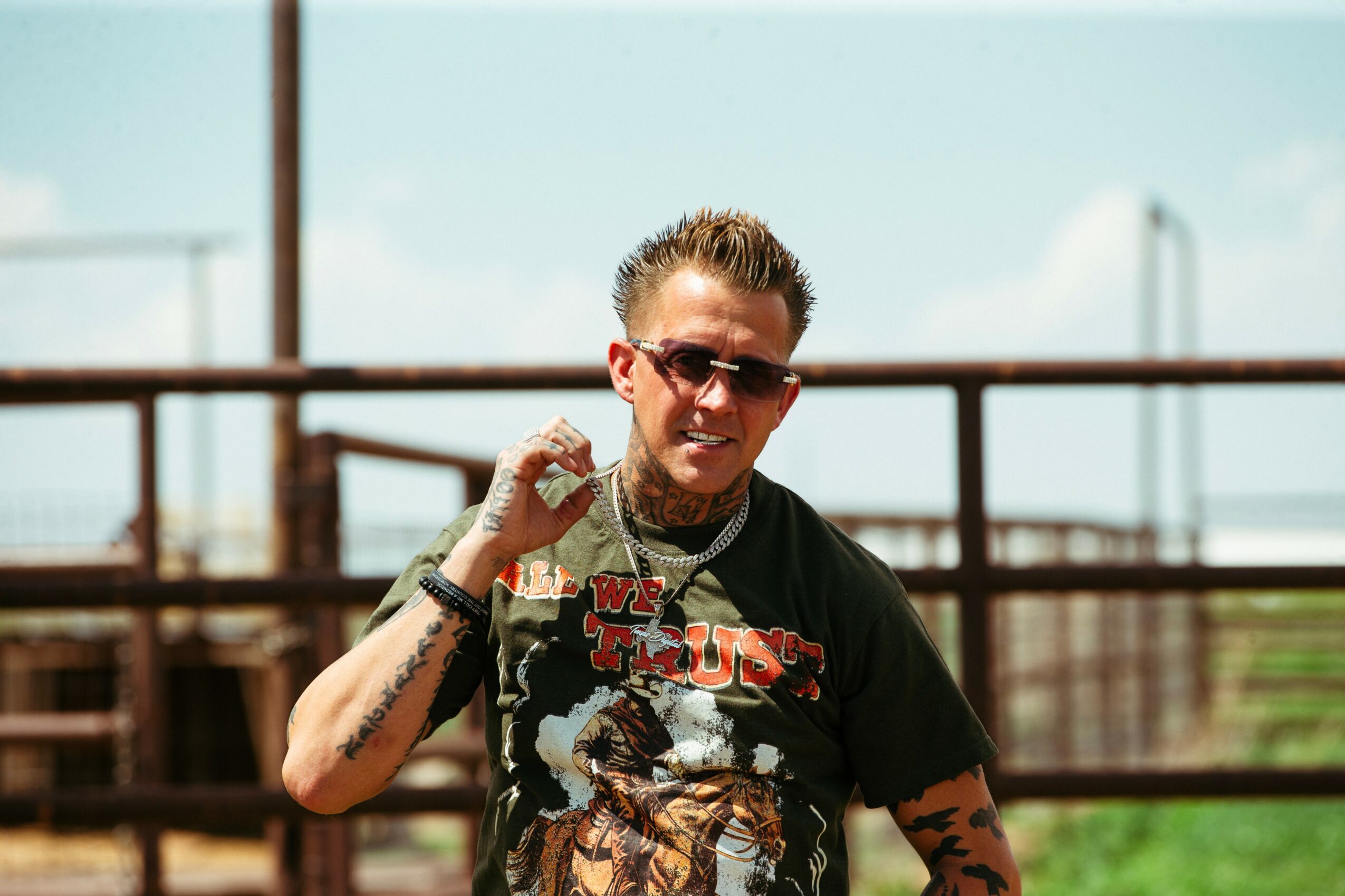 Man wearing sunglasses and a graphic T-shirt, smiling outdoors with a metal railing and blue sky in background.