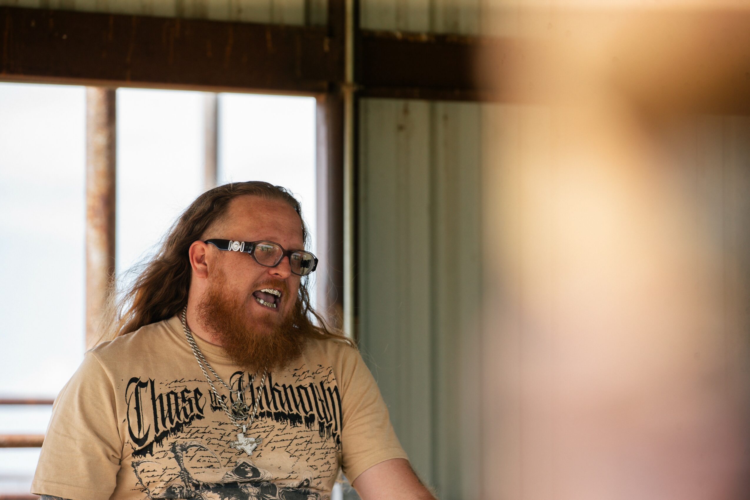 Person with long hair, glasses, and a beard smiling, wearing a beige T-shirt with text, indoors near a window.