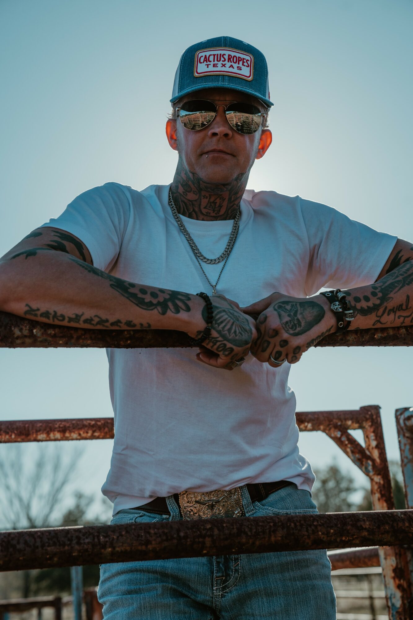 Young man with tattoos, wearing sunglasses, a cap, and a white t-shirt, leaning on a railing outdoors.