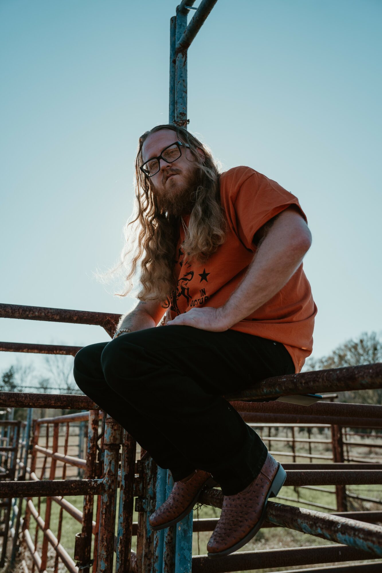 Person with long hair and glasses sitting on a metal railing outdoors, wearing an orange shirt and black pants.