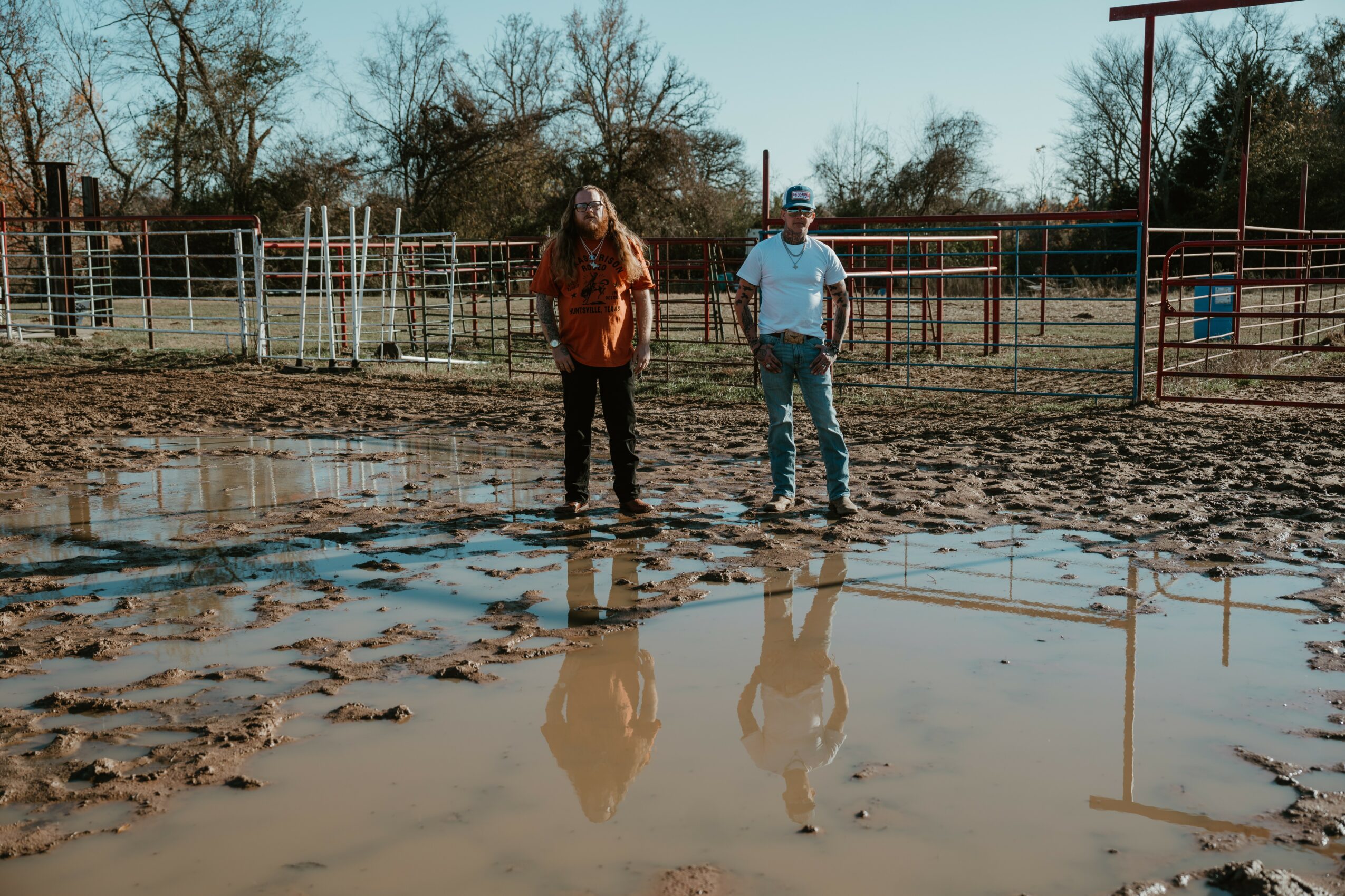 Two people standing on muddy ground with puddles, trees, and fences in the background, reflecting in the water.