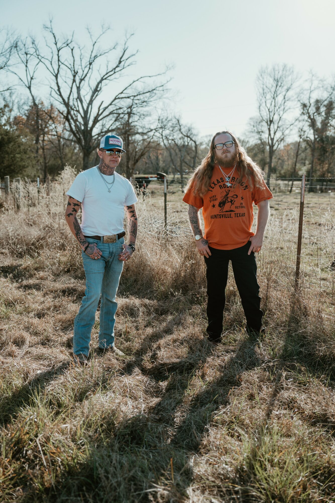 Two people standing outdoors in a grassy field with trees in the background, one wearing a white shirt and blue jeans, the other in an orange shirt and black pants.