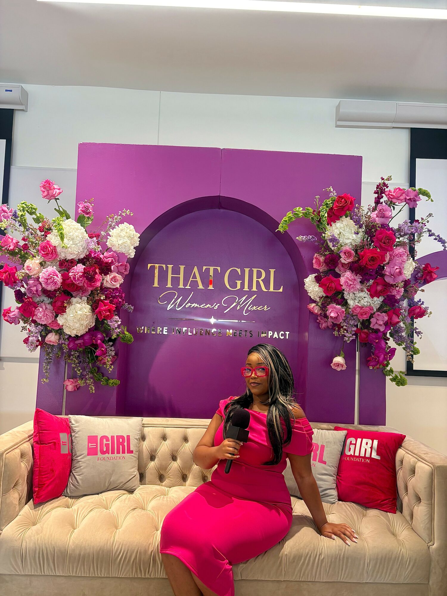 Woman in pink dress sitting on a beige sofa with pink pillows, floral arrangements, and a purple backdrop with text.