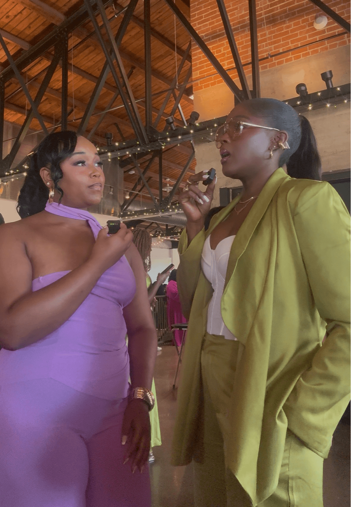 Two women in conversation indoors, one in a purple dress holding a glass, the other in a yellow suit with glasses.