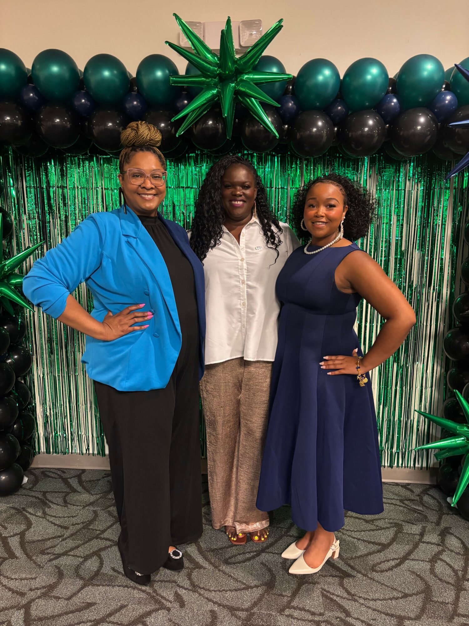 Three women standing together in front of green and black balloon decorations, smiling at the camera.