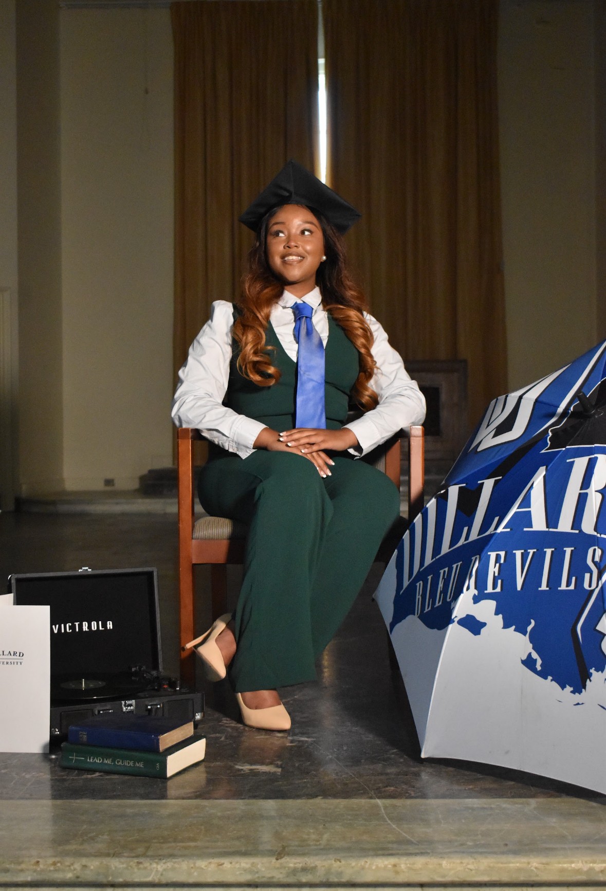 Young woman in graduation cap and gown seated on a chair, smiling, with a stage and banners in the background.