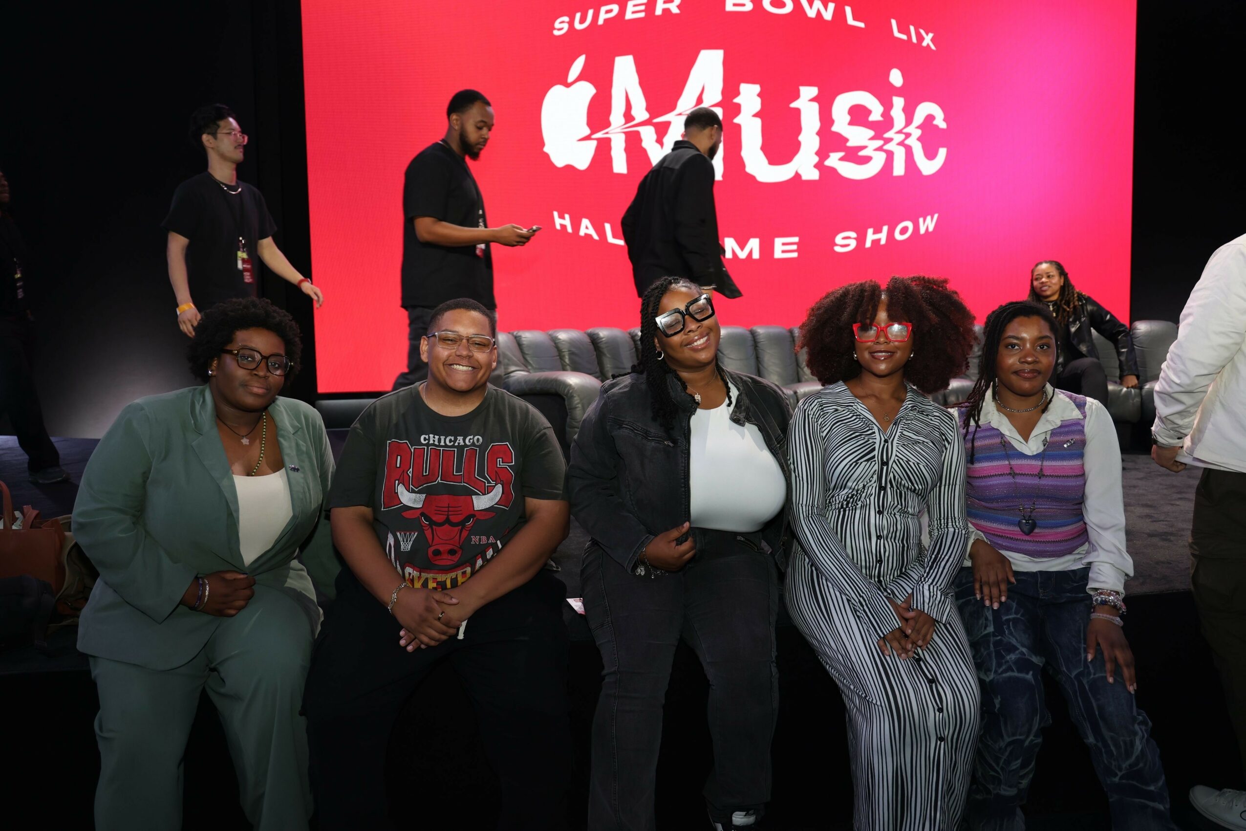 Group of six people standing in front of a pink Super Bowl music show backdrop, some smiling and posing for the photo.