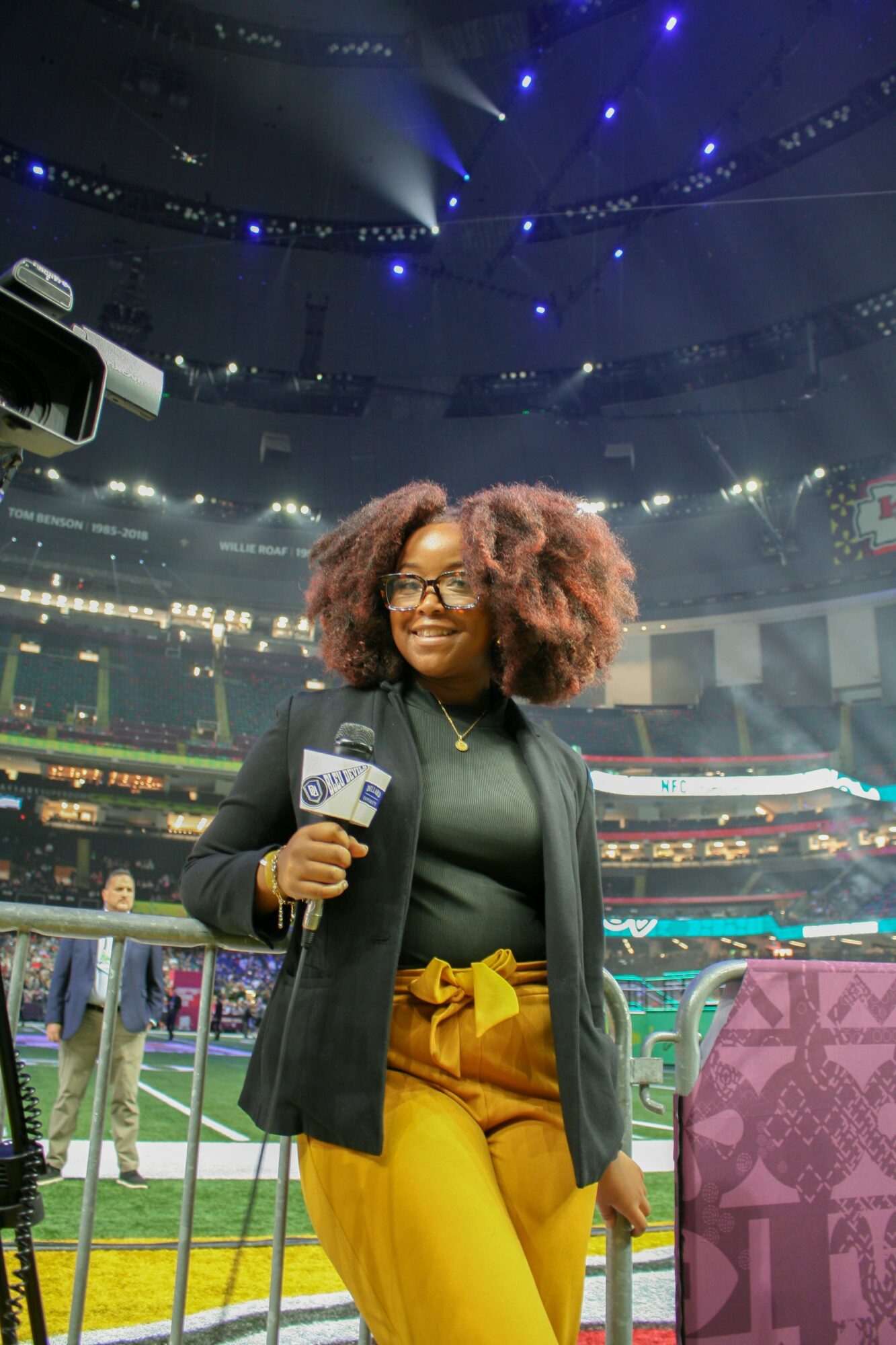 Woman with curly hair and glasses holding a microphone in a stadium with bright lights and a crowd.