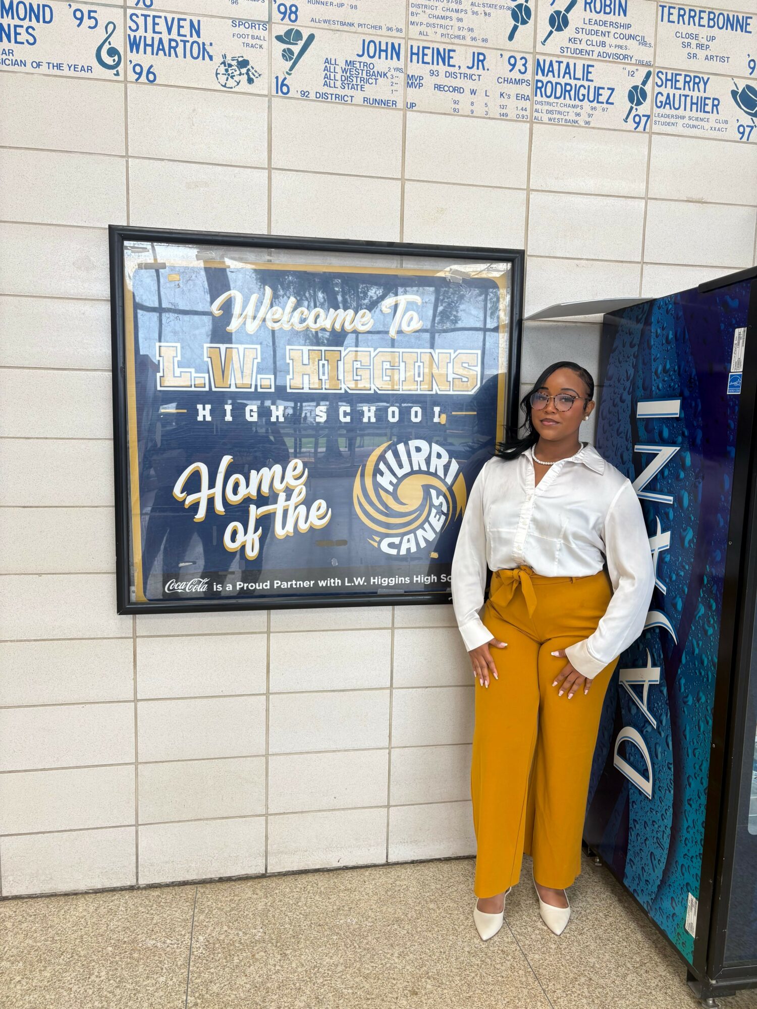 Woman standing next to a signboard with text, wearing a white top and yellow pants, leaning against a wall.