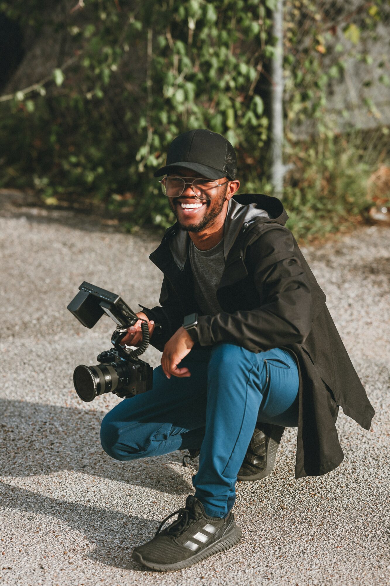 Person crouching outdoors holding a camera, smiling, wearing a black cap, glasses, black jacket, and blue pants.