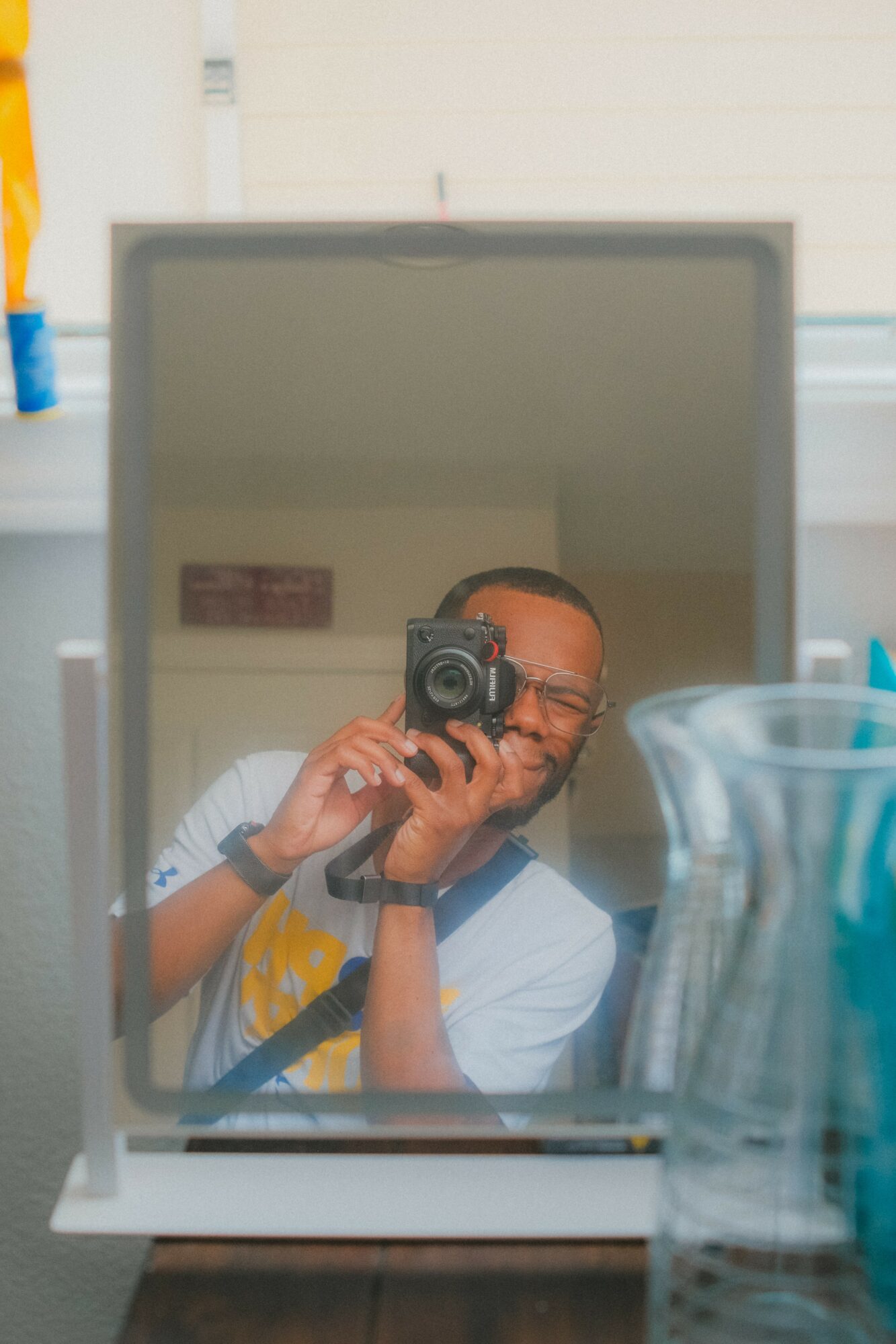 Person taking a photo with a camera, reflected in a mirror, with a blurred background and glassware in foreground.