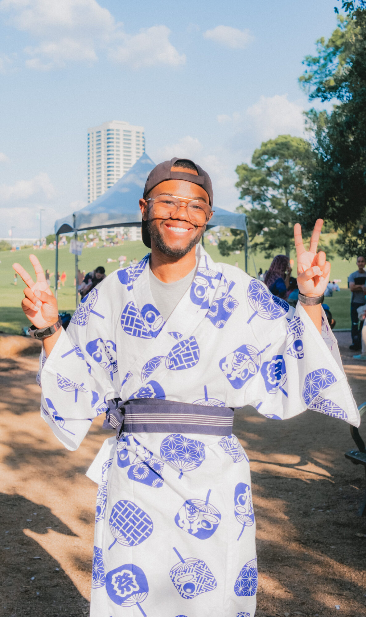 Smiling person in traditional Japanese yukata making peace signs outdoors, with trees and a building in background.