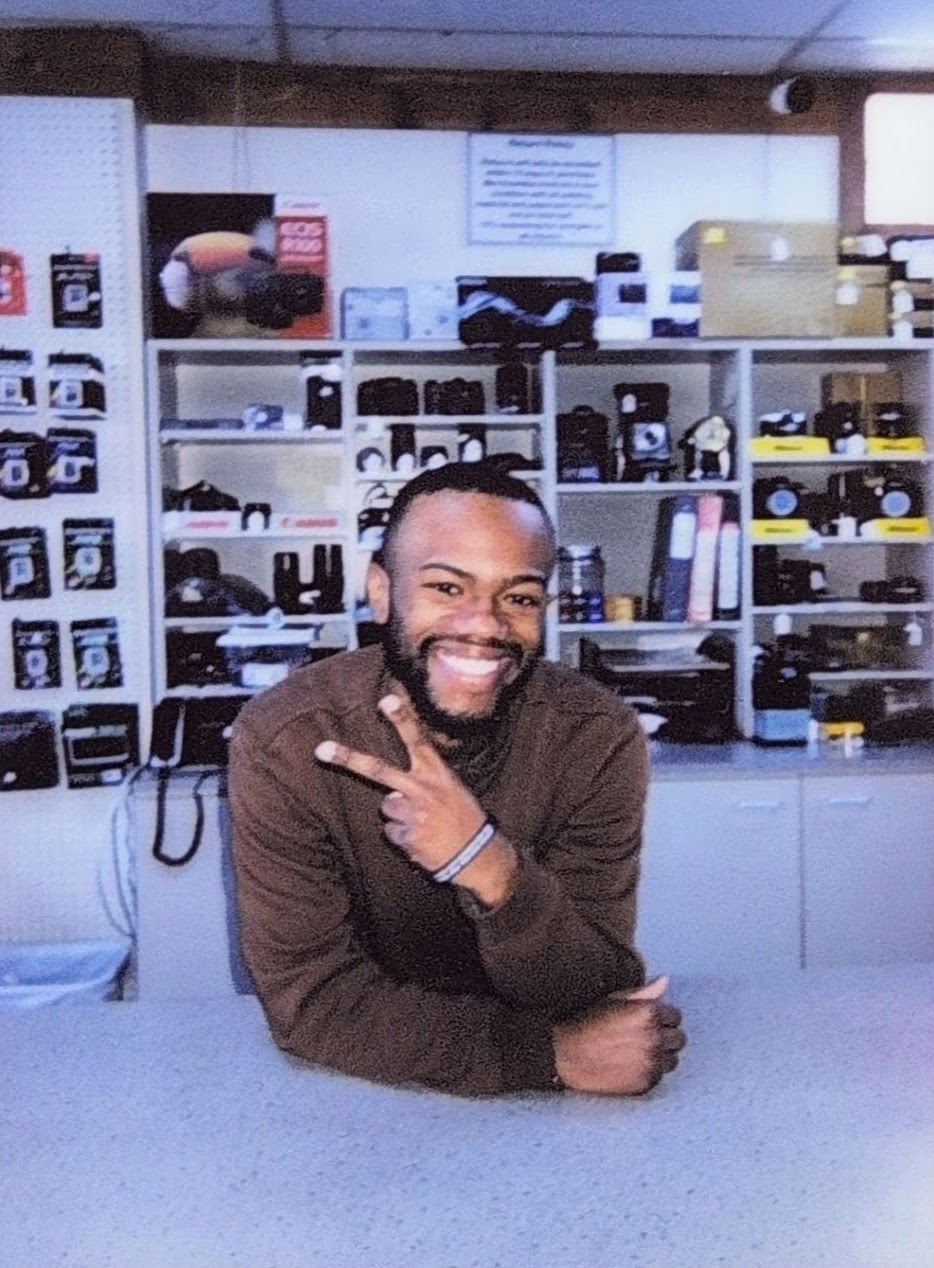 Smiling man with a beard making a peace sign in a camera store with shelves of cameras and accessories behind him.