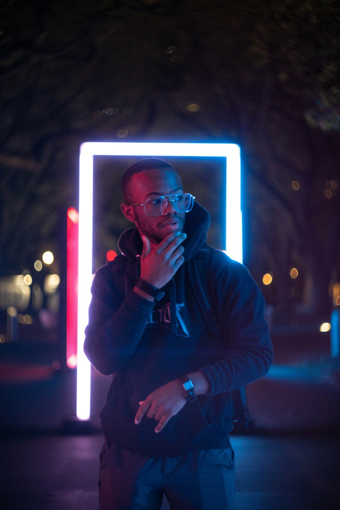Person wearing glasses and a hoodie, standing in front of illuminated rectangular frame at night, hand near face, city lights in background.