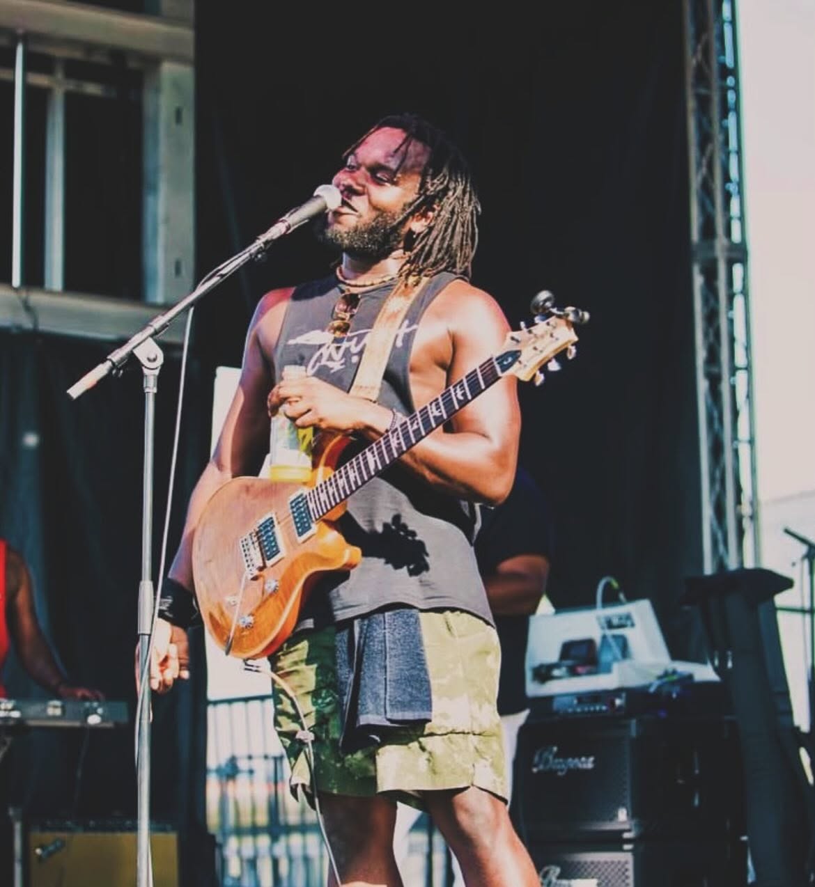Person with dreadlocks singing into a microphone while holding an electric guitar on stage outdoors.