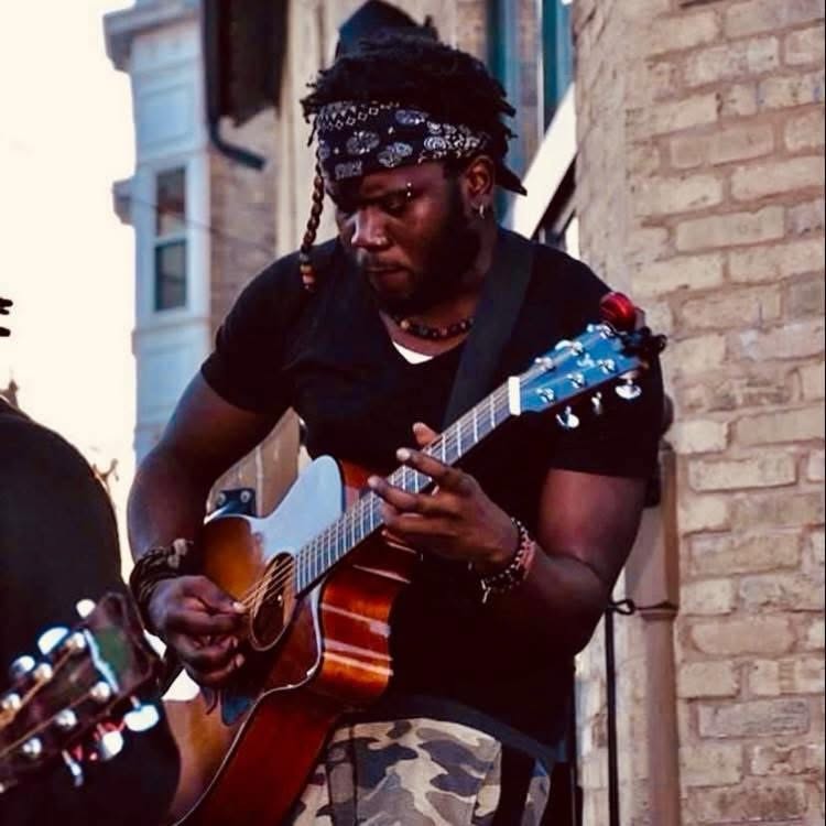 Person playing guitar outdoors, wearing a black shirt and bandana, with a brick building in the background.