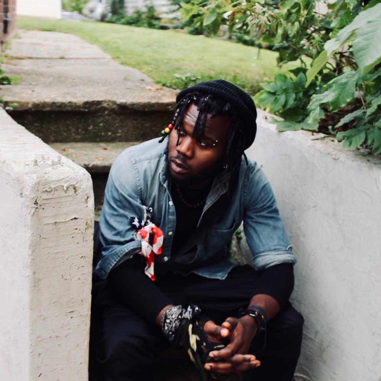 Young man with braided hair, wearing a denim jacket and black clothing, sitting outdoors among greenery.