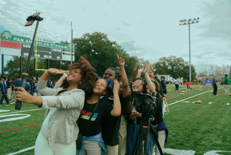 Group of people taking a selfie on a sports field with a scoreboard and trees in the background.