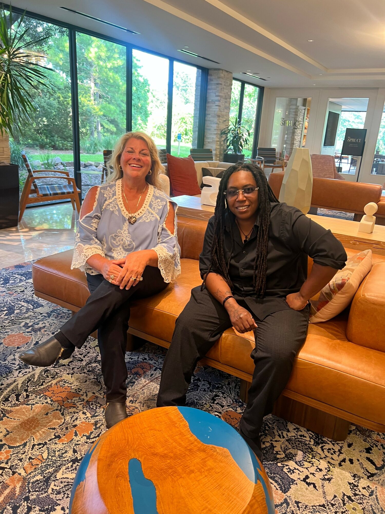 Two women sitting on a sofa in a bright room with large windows and greenery outside, smiling and talking.