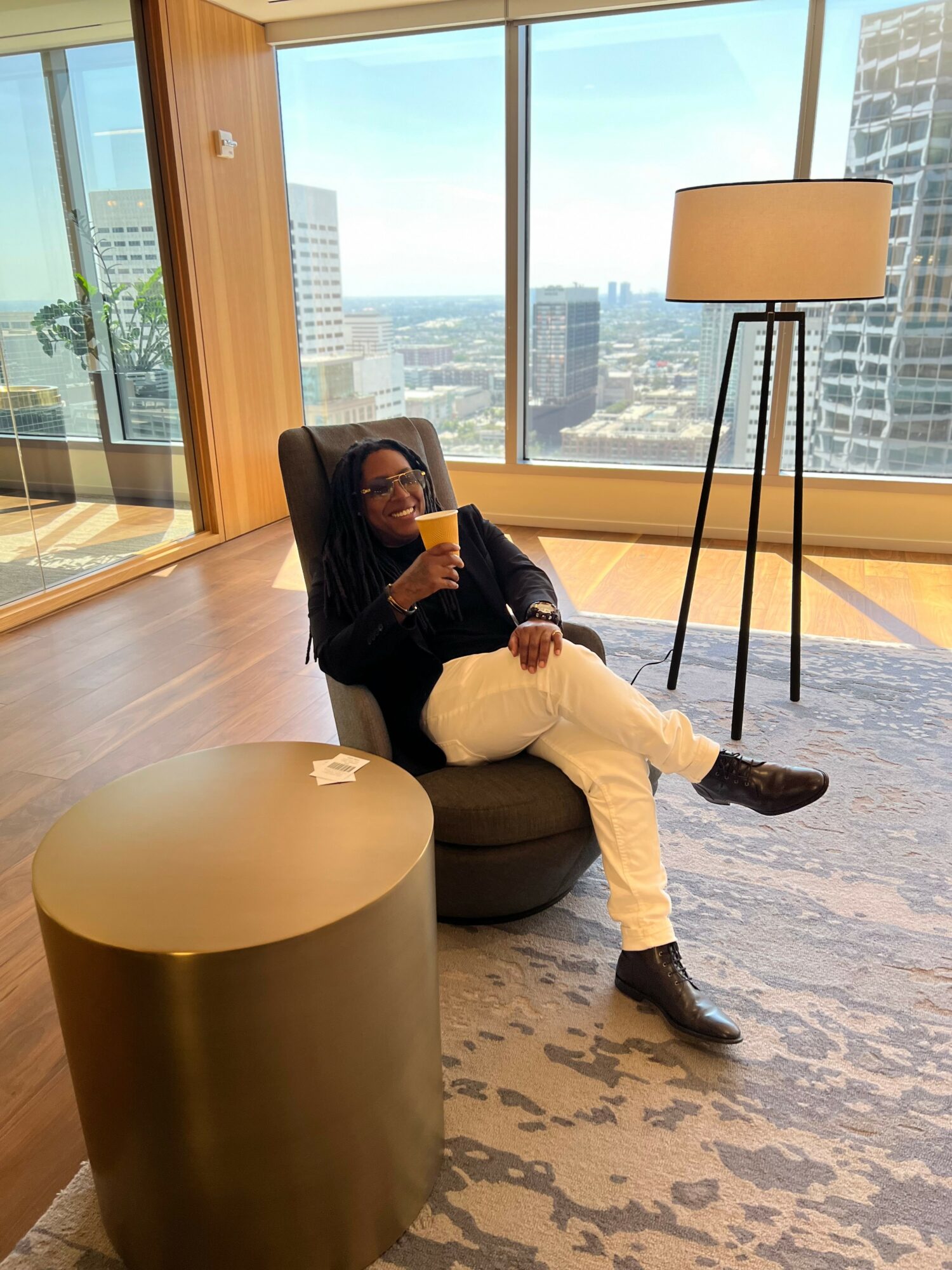 Person sitting in a modern room with large windows, holding a yellow drink, with cityscape view outside.