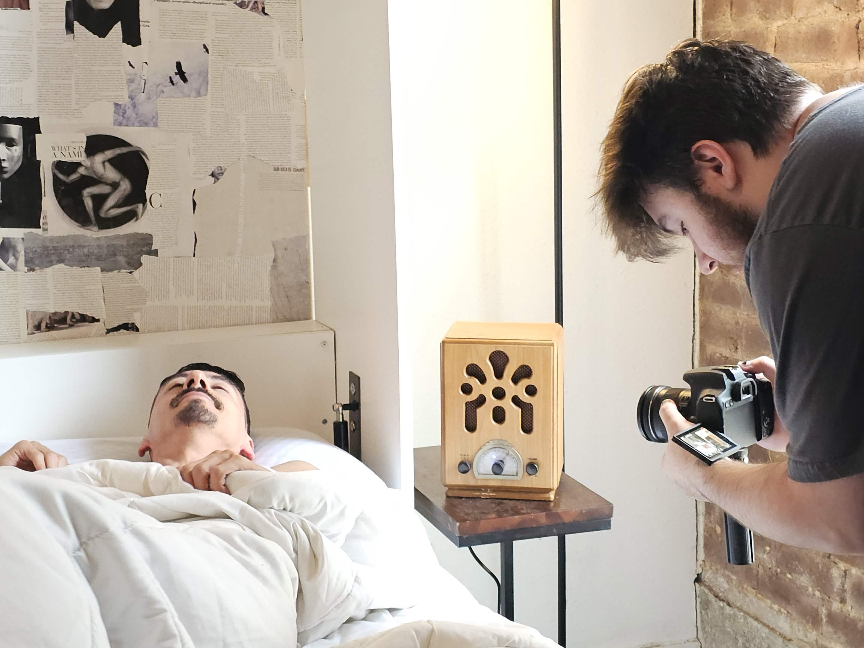 Person lying in bed with eyes closed, another person taking a photo nearby, room with wall art and a speaker on a table.